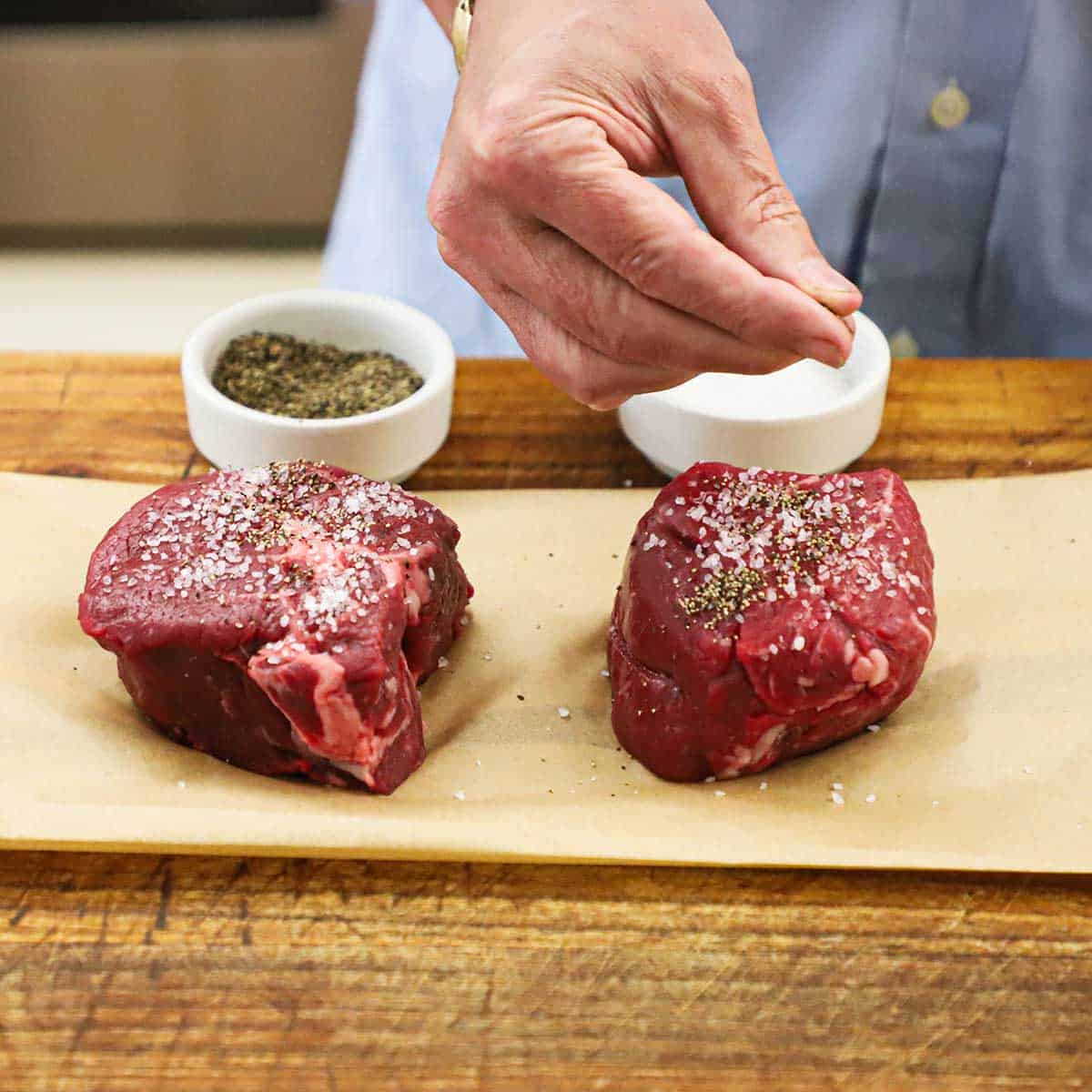 A person using his fingers to sprinkle ground black pepper over the tops of two uncooked filet mignon steaks on brown paper.