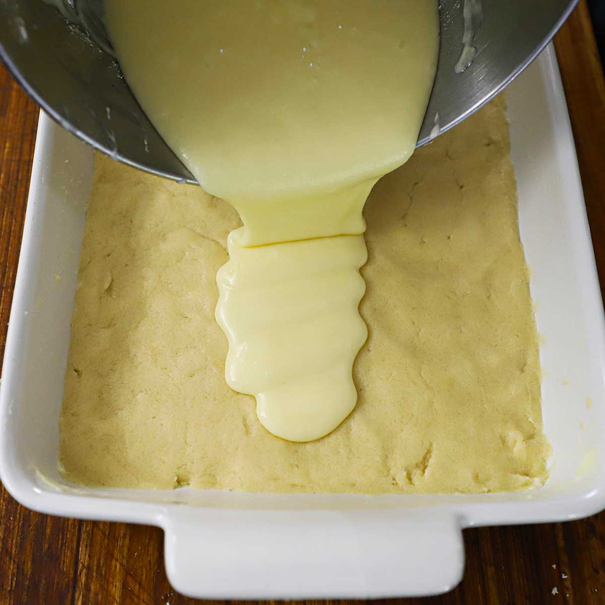 A person pouring a cream cheese mixture over an uncooked crust in a white 9 by 13 inch baking dish.