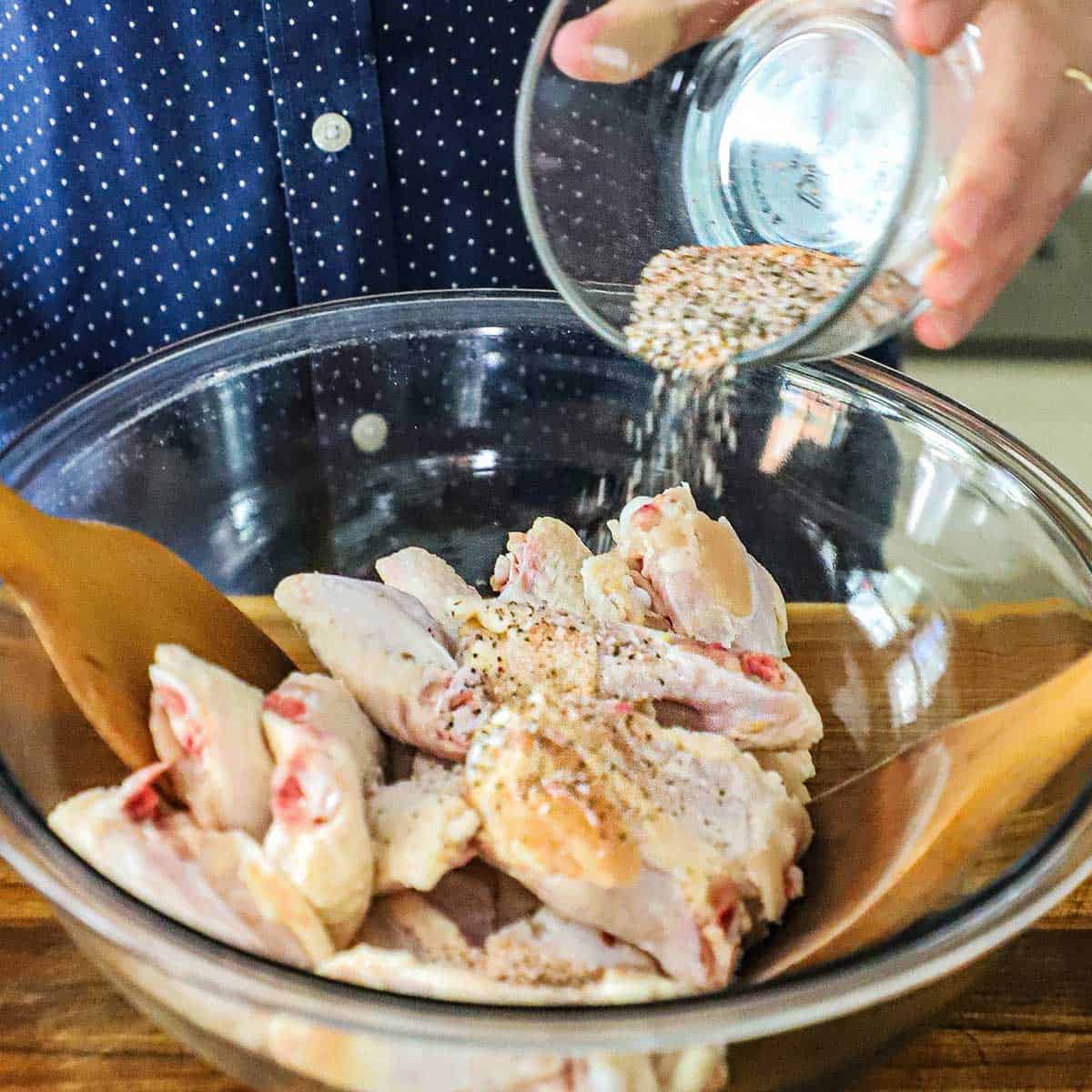 A person transferring a mixture of seasonings over uncooked chicken wings in a large glass bowl.
