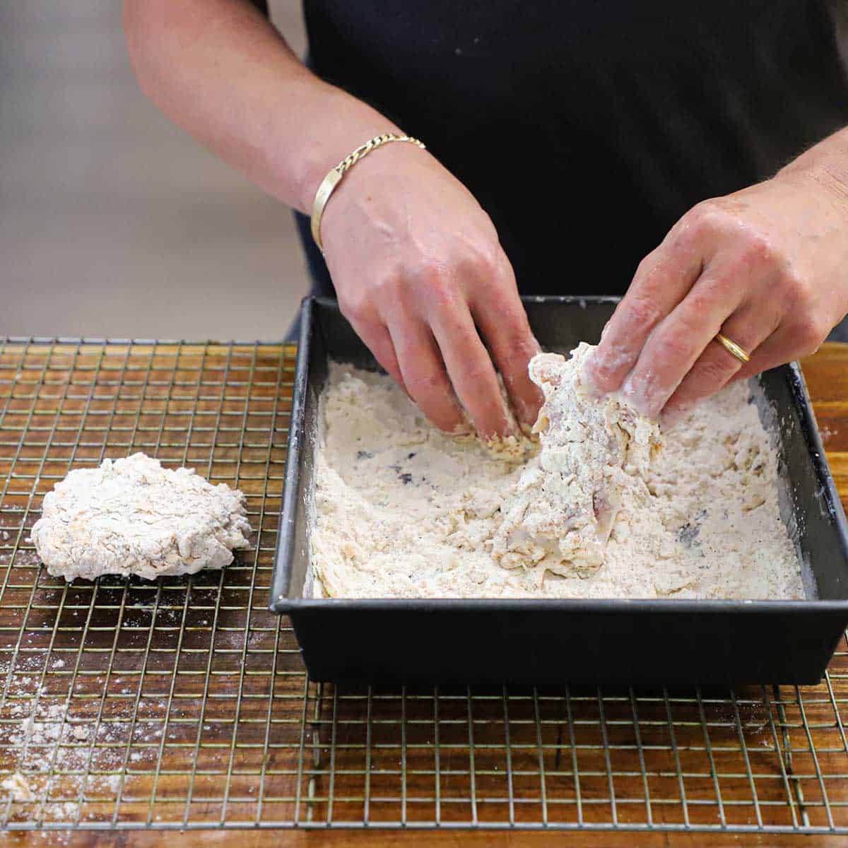 A person dredging chicken thighs that have been coated with seasoned buttermilk in a square pan filled with seasoned flour.