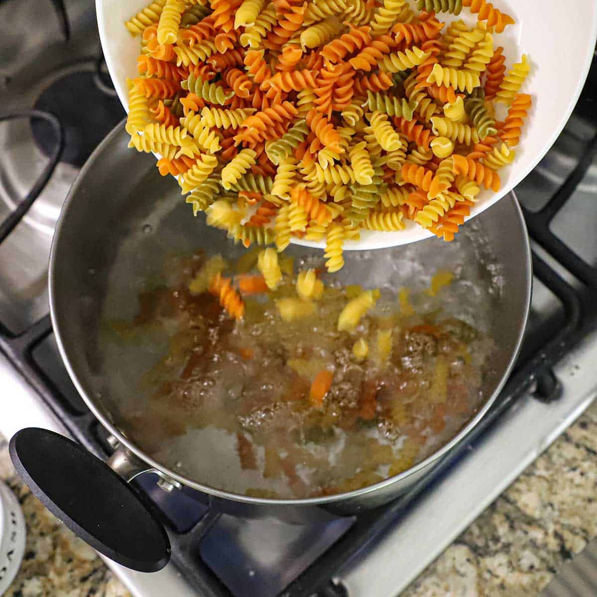 A person transferring uncooked tricolor rotini pasta into a pot of boiling water on a gas stove.