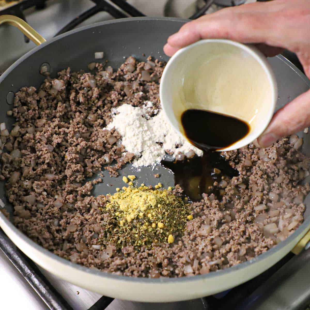 A person pouring Worcestershire sauce from a small white bowl into a skillet that is filled with cooked ground beef, onions, and small piles of flour, dried herbs, and dry mustard.