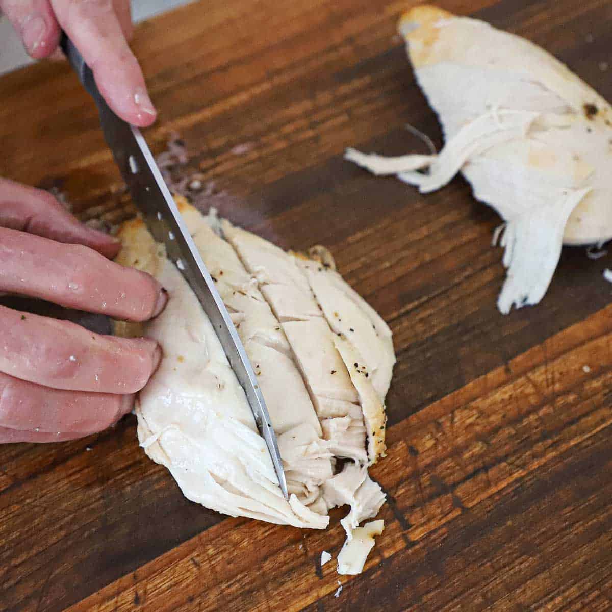A person using a chef's knife to cube a cooked piece of chicken breast on a large cutting board.