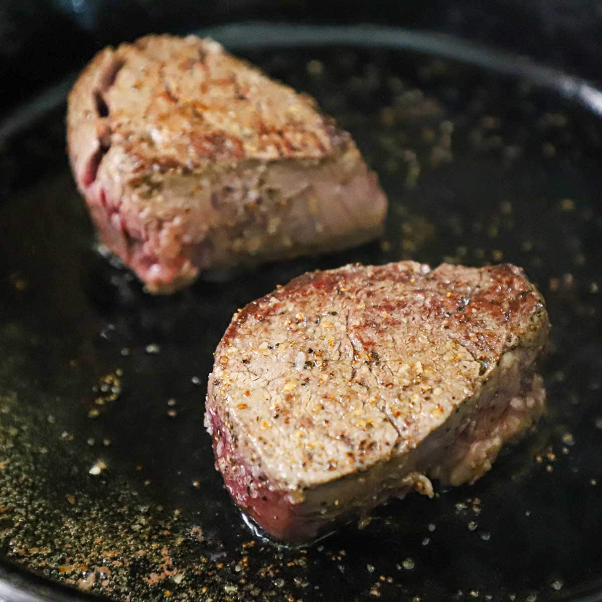 Two large filet mignon steaks being seared in a large black cast-iron skillet filled with shimmering melted lard.