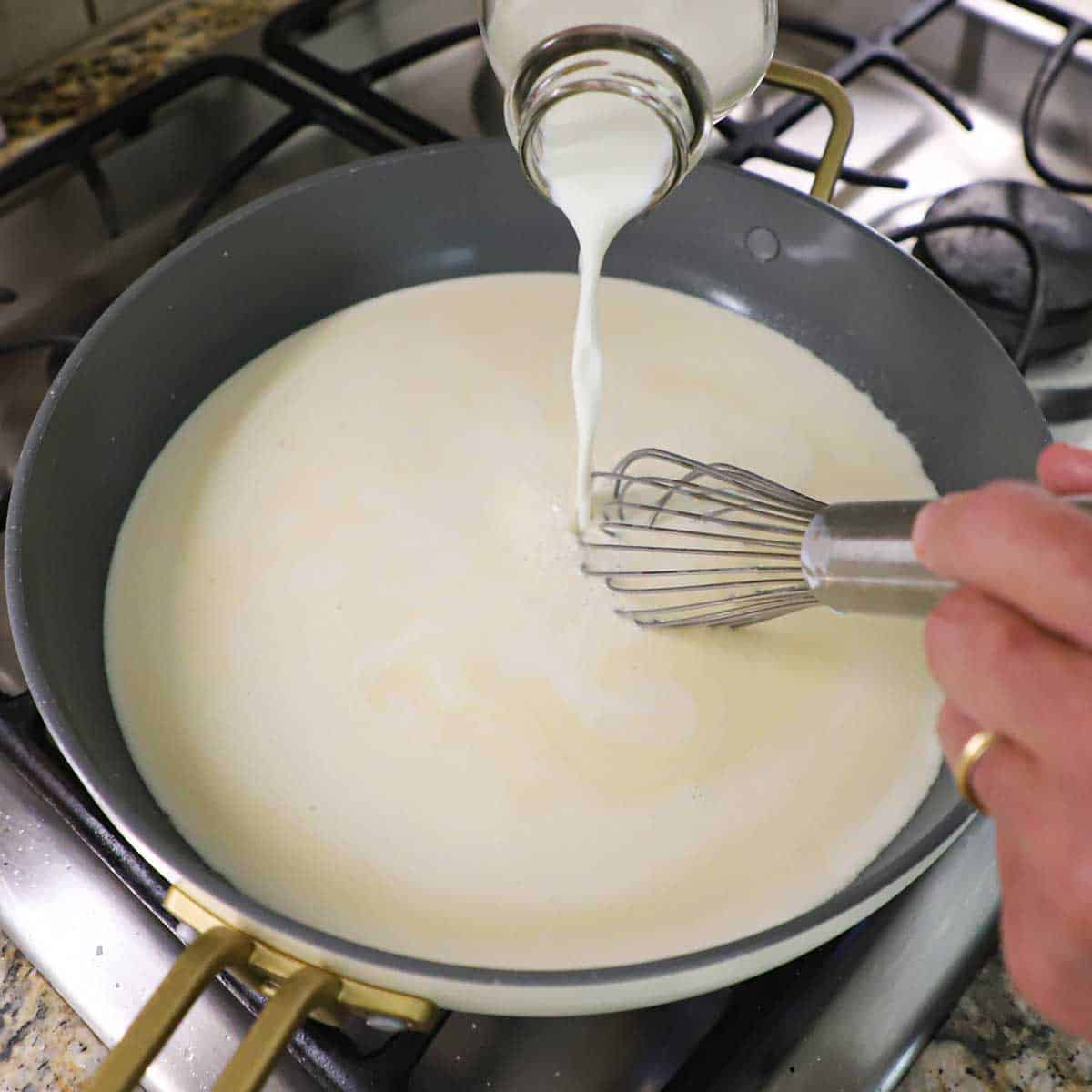 A person pouring milk with one hand into a skillet filled with a bechamel sauce with his other hand whisking the mixture.