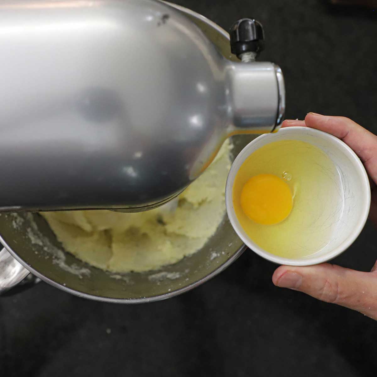 A person holding a small white bowl that is filled with a whole egg over a stand mixer filled with creamed butter and sugar.