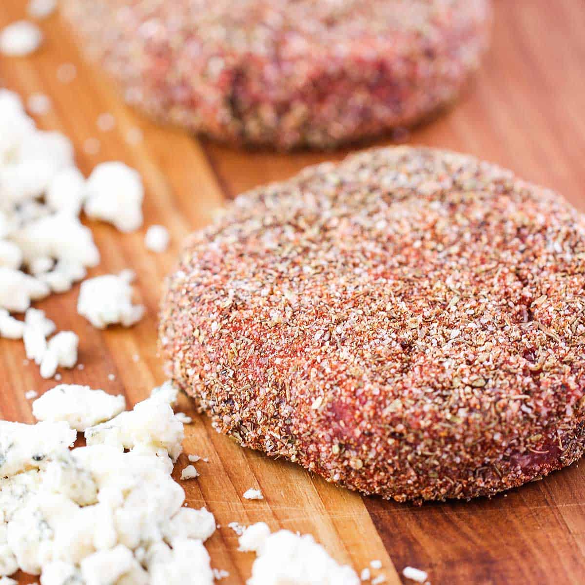 Two uncooked beef patties that are coated with a blackening seasoning on a wooden cutting board next to crumbled blue cheese.