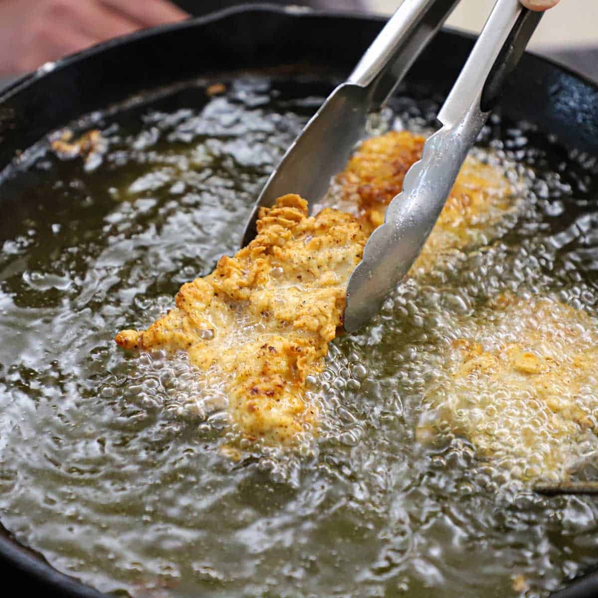 A person using a pair of metal tongs to grip onto a strip of a breaded chicken thigh that is being fried in hot oil in a black cast-iron skillet.