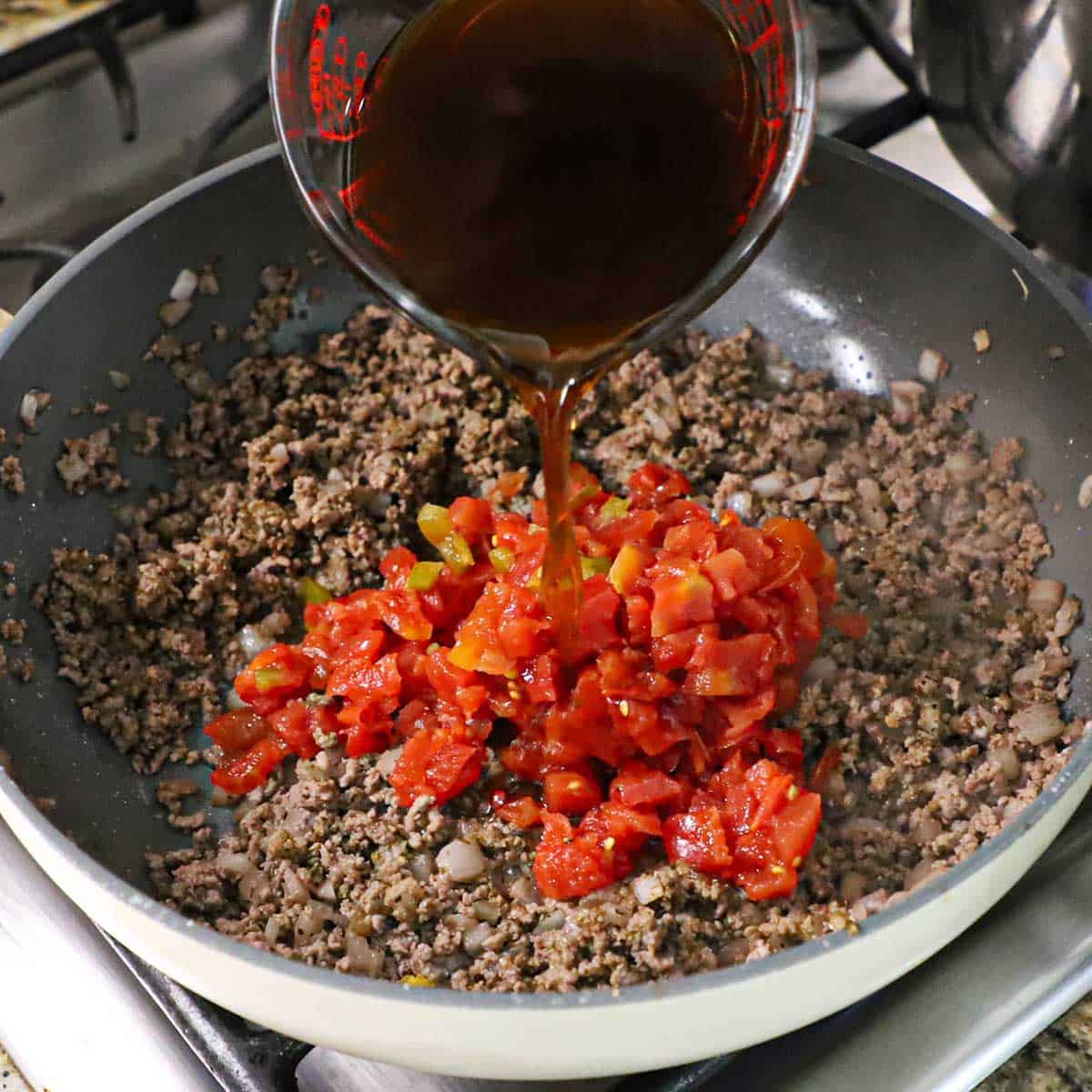 Beef stock being poured from a glass measuring cup into a skillet that is filled with seasoned cooked ground beef and a pile of chopped tomatoes.