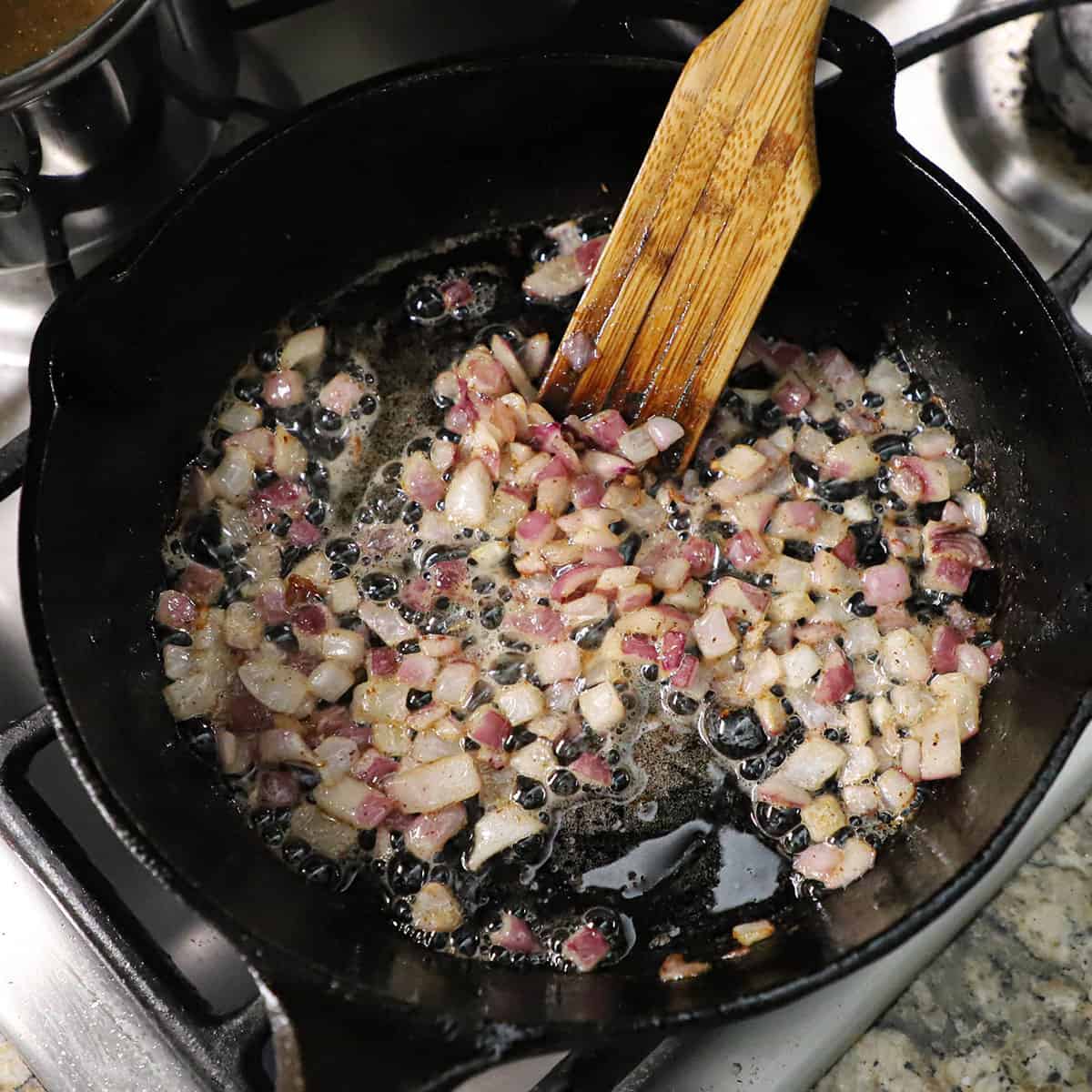 A person using a wooden spatula to stir chopped bacon and red onion that are sizzling and being cooked in a black cast-iron skillet.