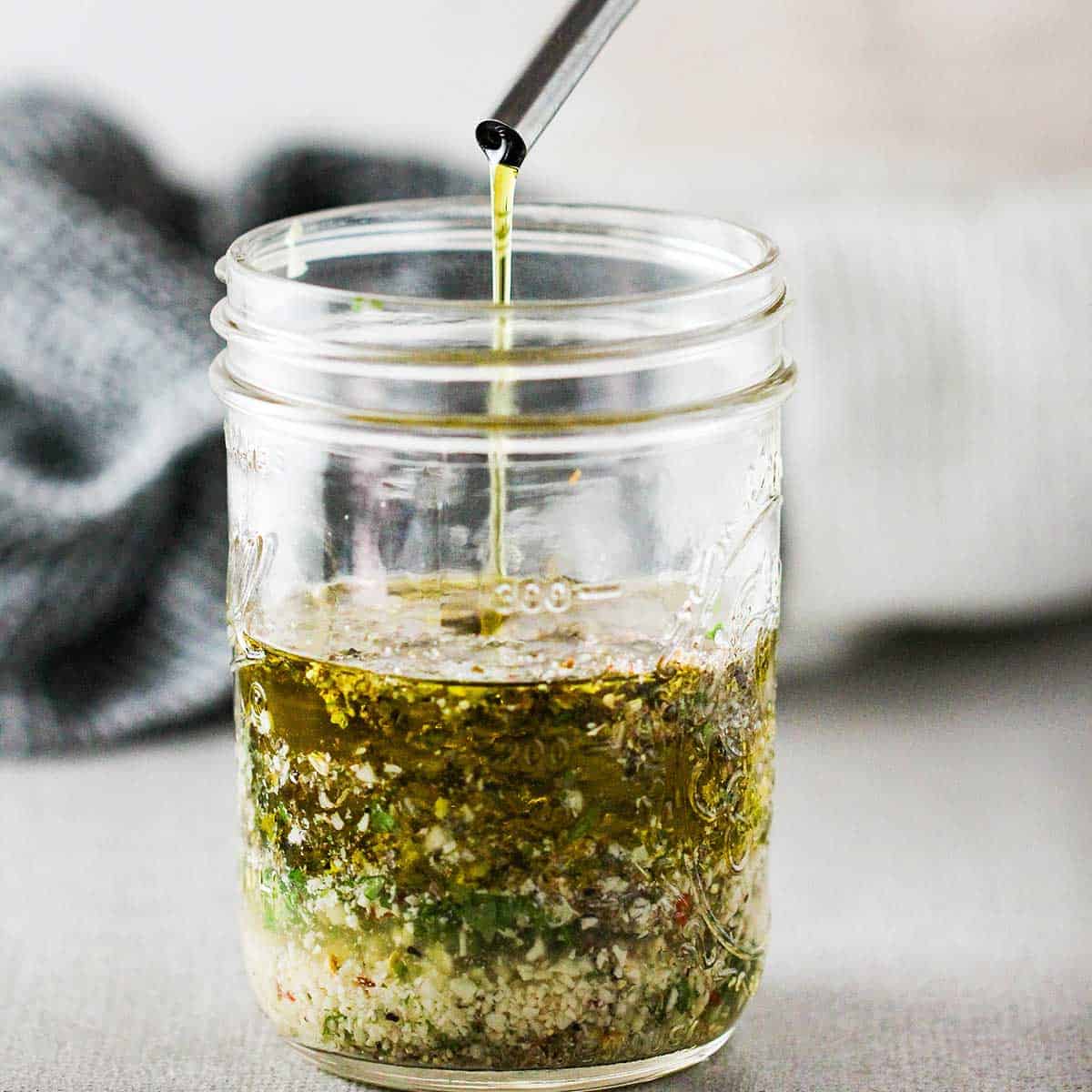 A person pouring extra-virgin olive oil from a small tube into a glass jar that is filled with the ingredients for homemade Italian dressing.