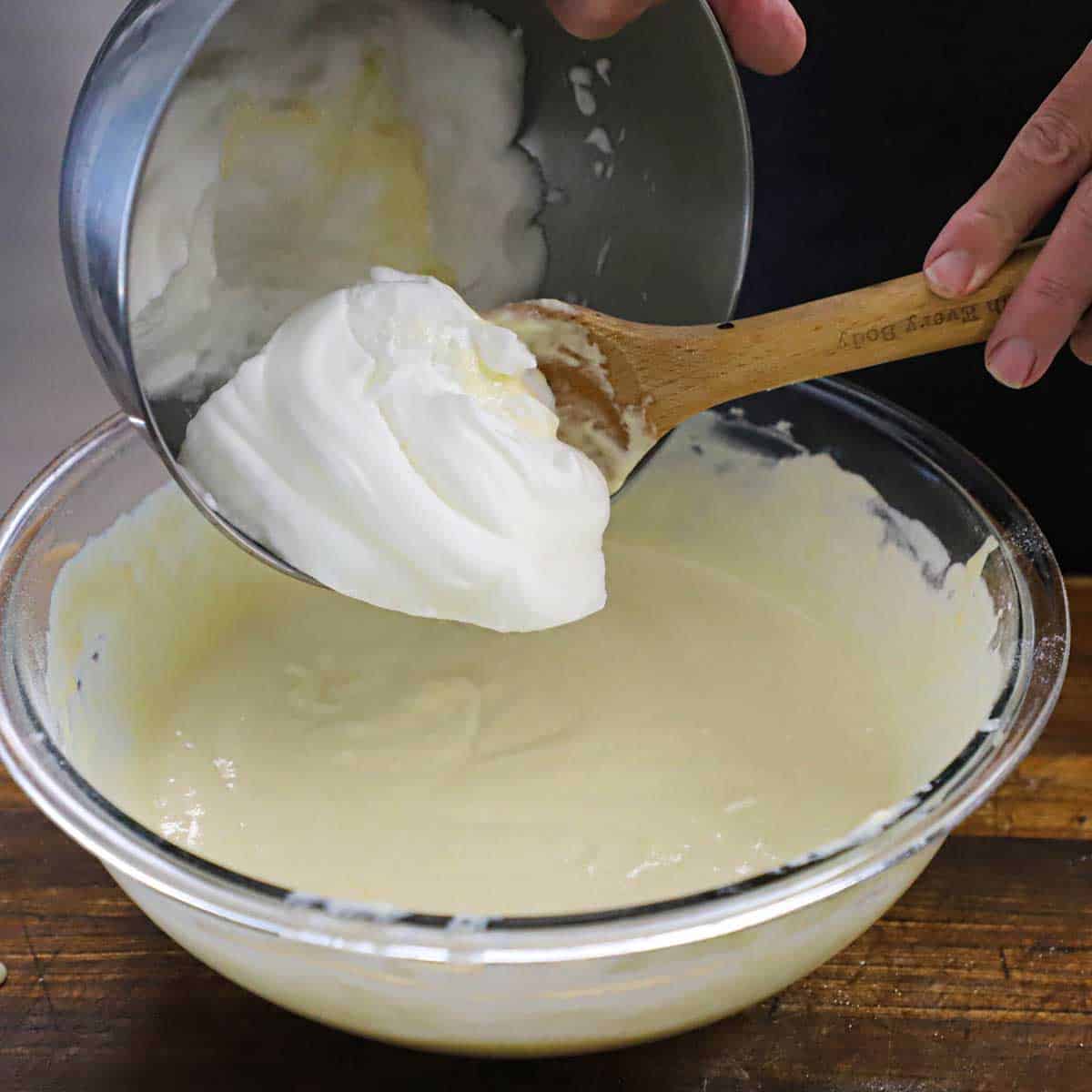 A person transferring soft peak egg whites from a metal bowl into a large glass bowl that is filled with buttermilk waffles batter.