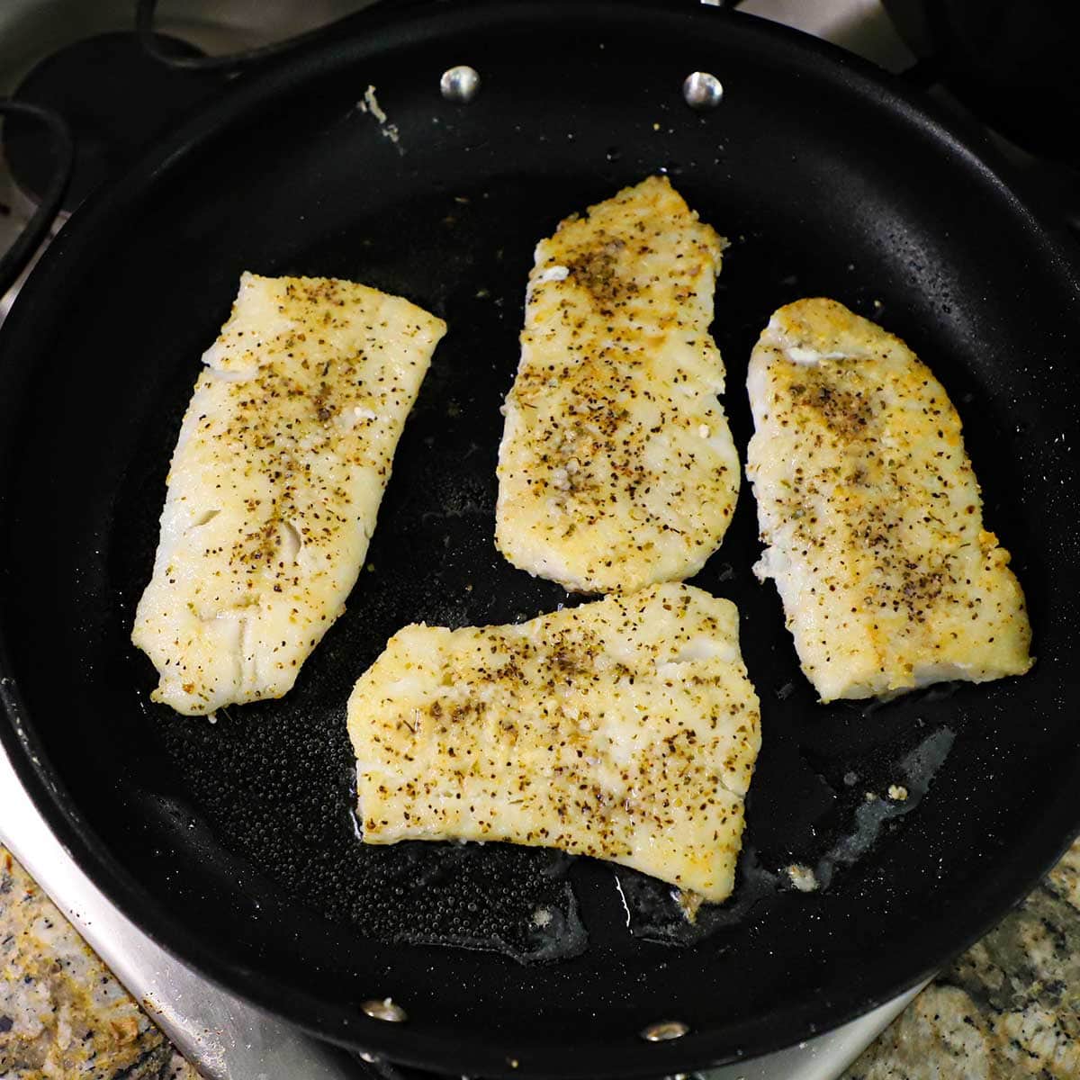 Four filets of cod being pan fried in olive oil and melted butter in a black non-stick skillet on a gas stove.