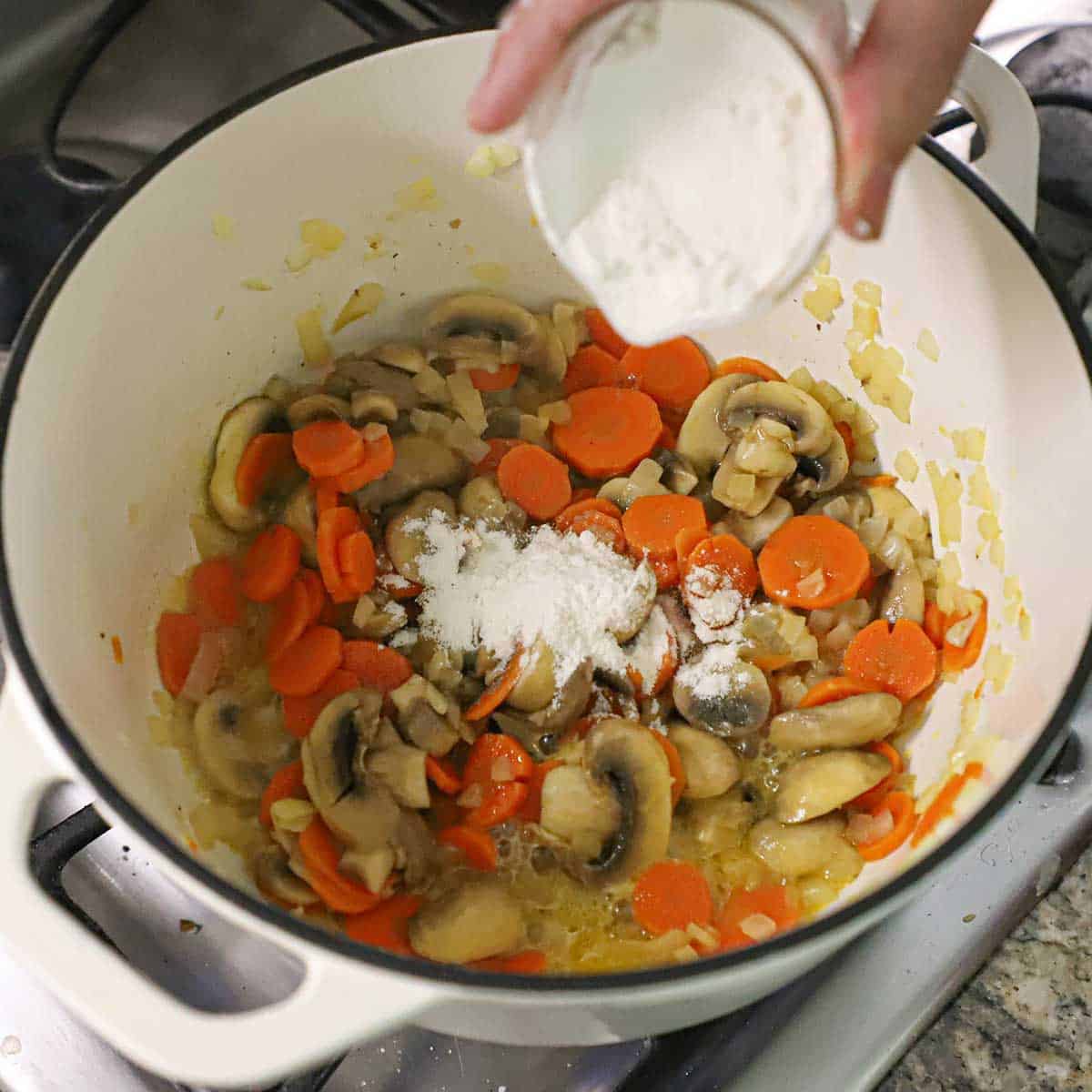 A person dumping all-purpose flour from a small white bowl into a pot that is filled with sautéed onions, carrot slices, and chopped onions.