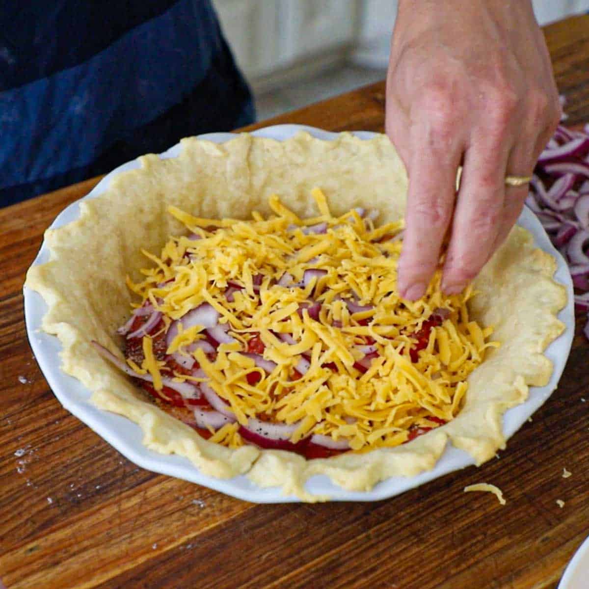 A person sprinkling shredded cheddar cheese over a layer of sliced tomatoes and slivers of red onion all in a partially pie crust in a pie dish.