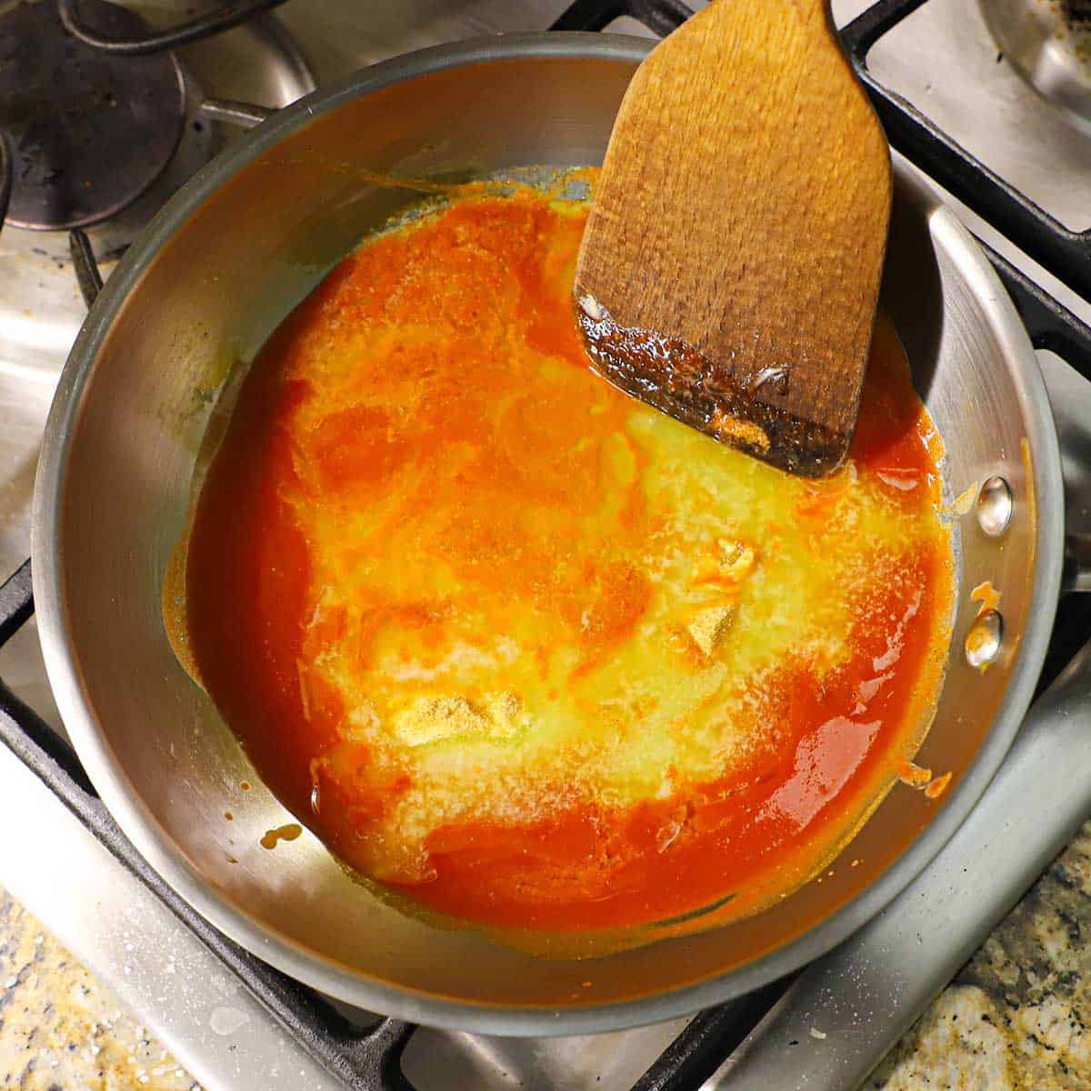 A person using a wooden spatula to stir melted butter in a saucepan filled with simmering hot sauce and a sprinkle of garlic powder.