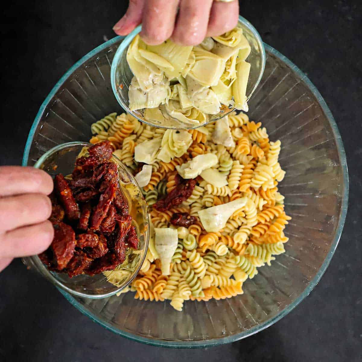 A person using one hand to transfer of marinated artichoke hearts into a bowl of cooked pasta and his other hand transferring pieces of sun-dried tomatoes into the same bowl.