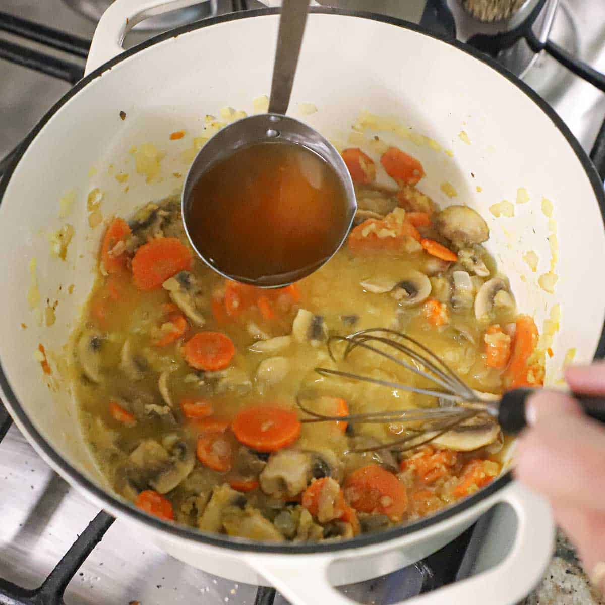 A person using a ladle to transfer chicken broth into a pot filled with sautéd vegetables that have been cooked with flour to create a roux.