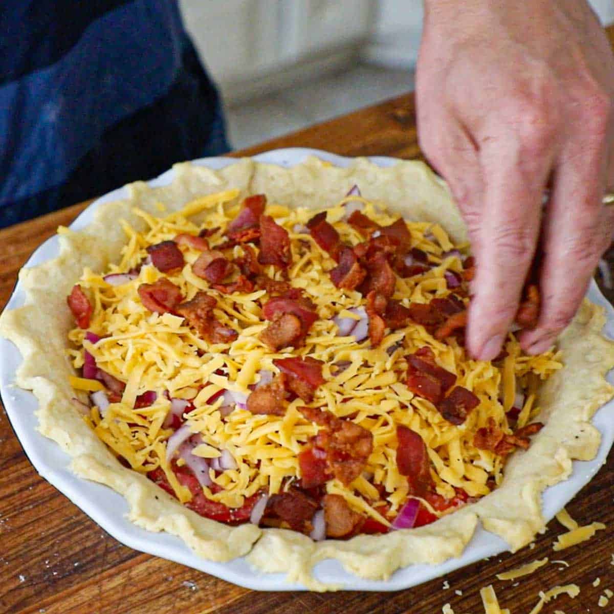 A person sprinkling crispy, cooked pieces of bacon over the top of a tomato cheddar and bacon pie resting on a wooden cutting board.