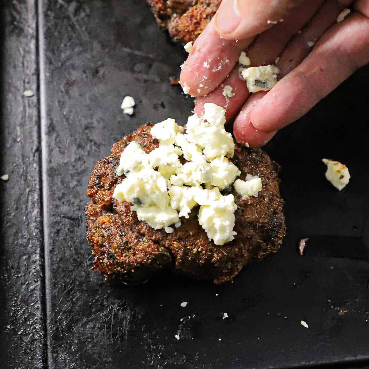 A person placing crumbled blue cheese on top of a cooked blackened hamburger patty.