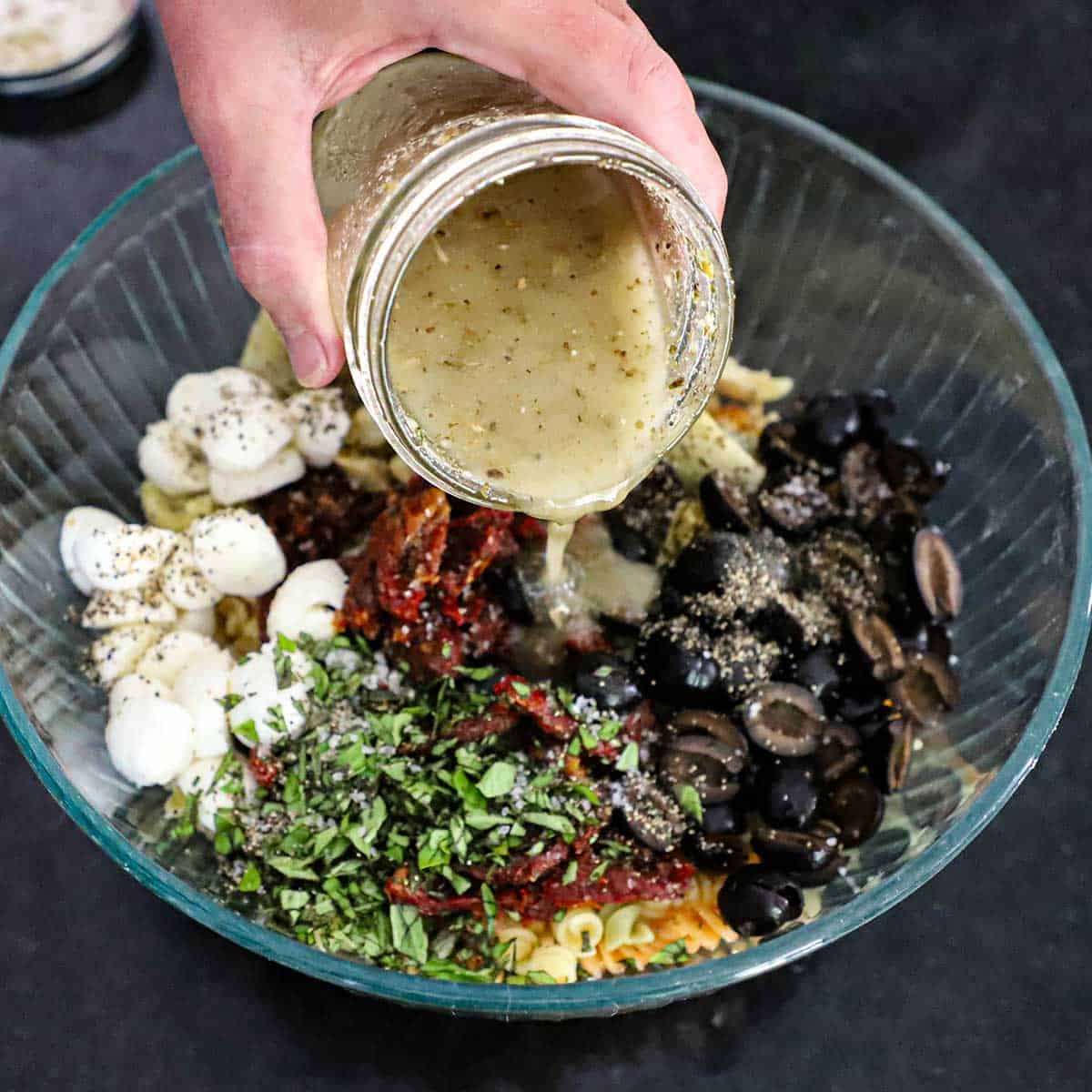 A person pouring homemade Italian dressing from a glass jar into a glass bowl filled with cooked rotini pasta, mozzarella balls, chopped basil, artichoke hearts, and halved black olives.