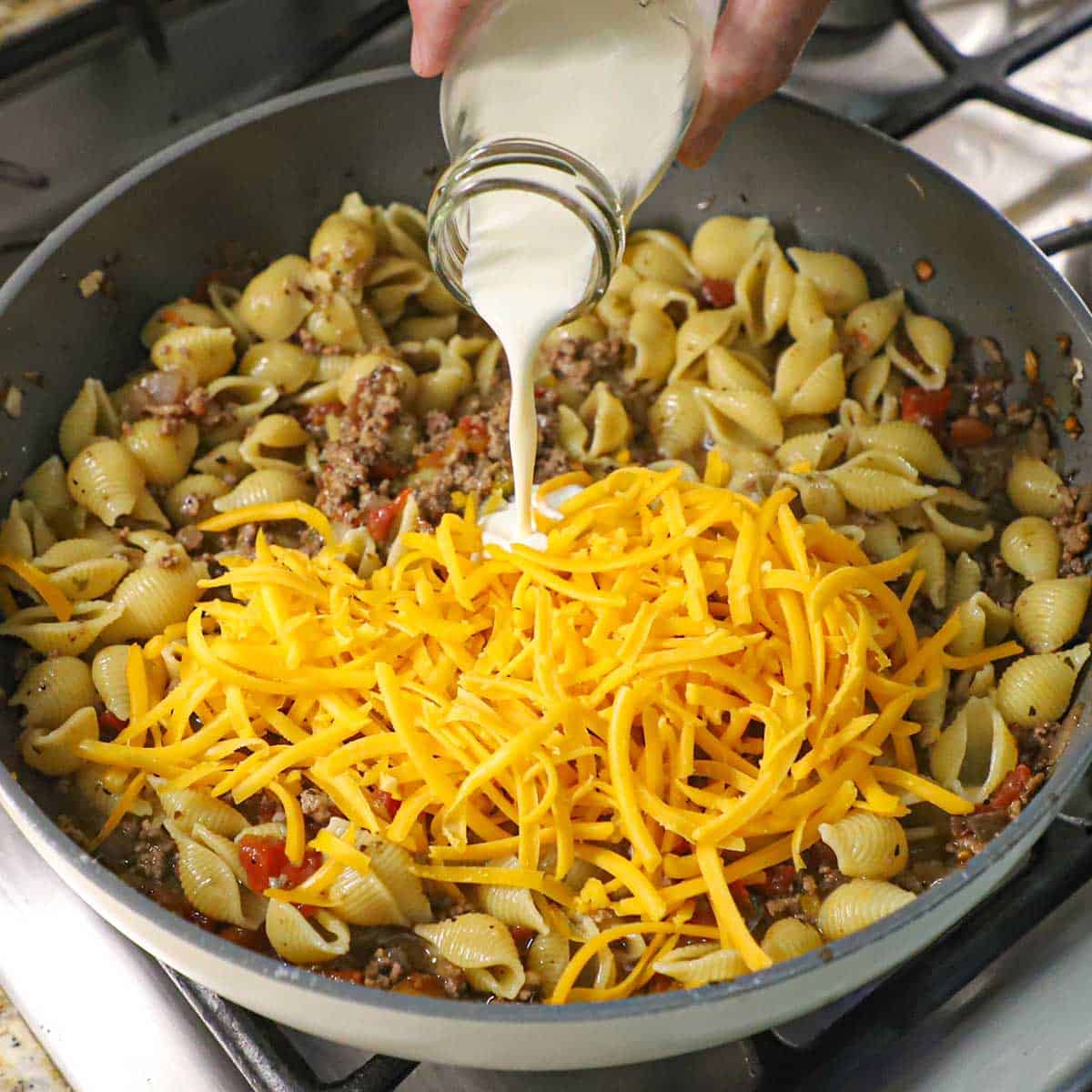 A person pouring heavy cream from a small glass milk jug into a skillet filled with pasta shells in meat sauce and topped with a pile of shredded cheddar cheese.