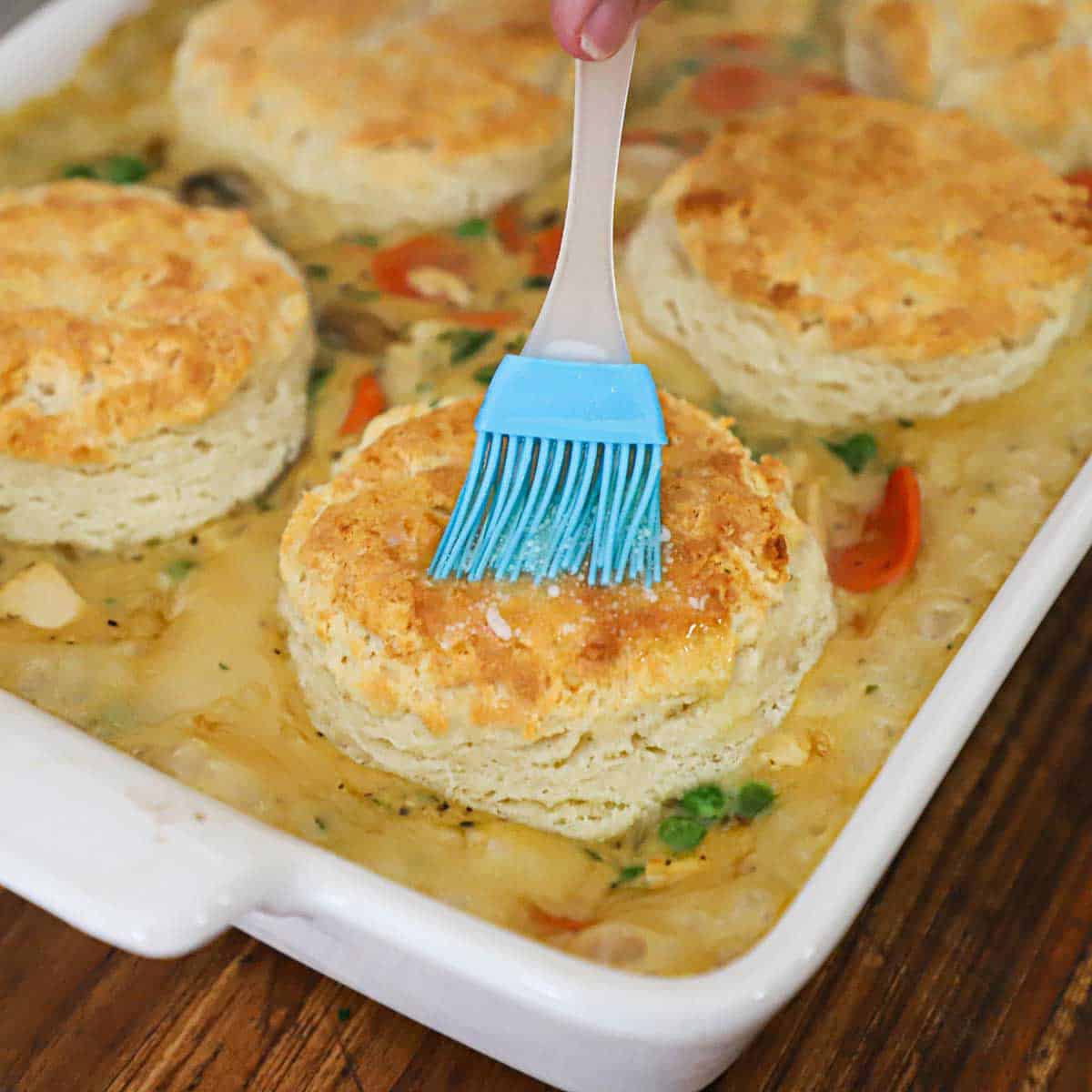 A person using a blue pastry brush to apply melted butter over the top of a biscuit that in a freshly baked chicken and biscuits casserole.