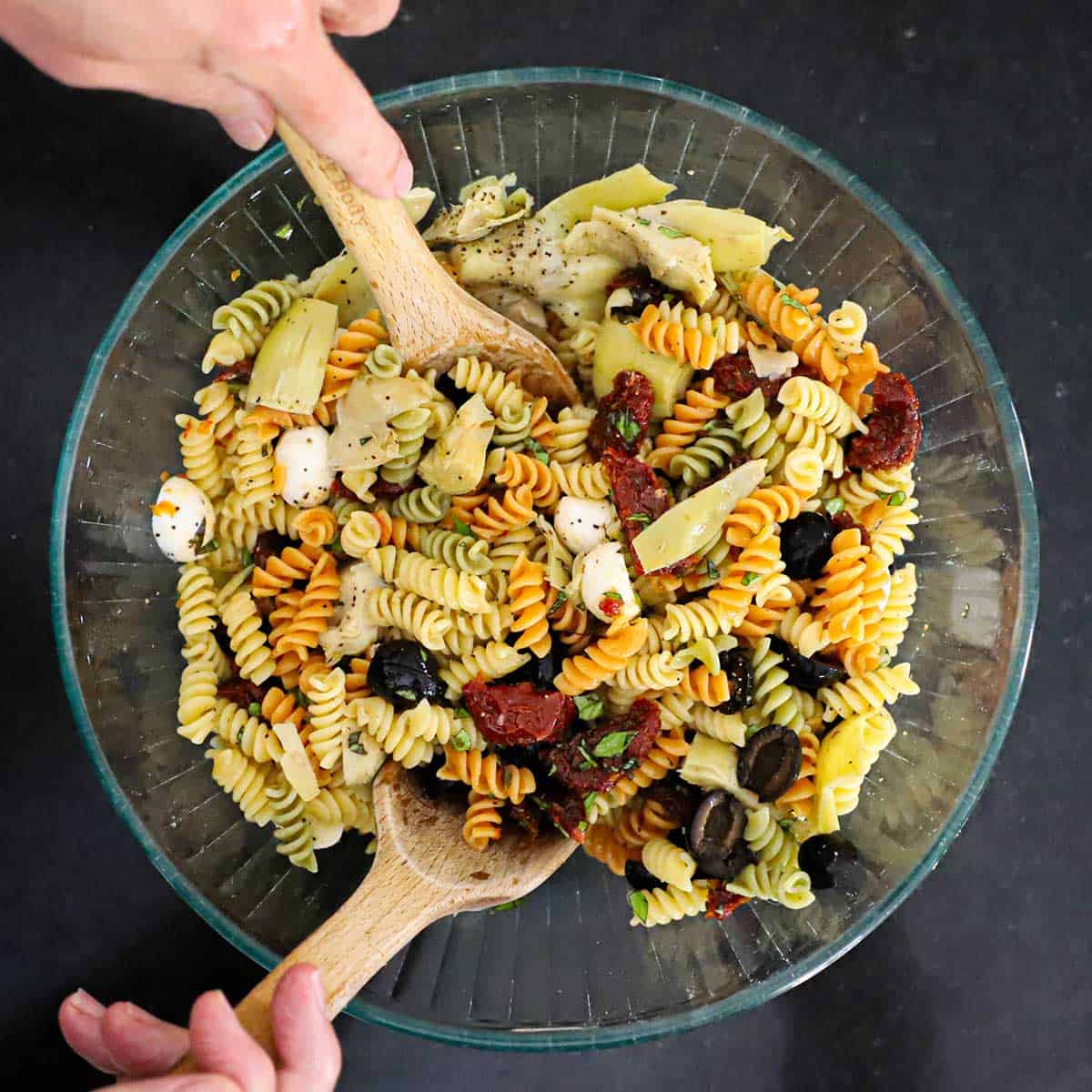 A person using two large wooden spoons to toss together a classic pasta salad in a large glass bowl.