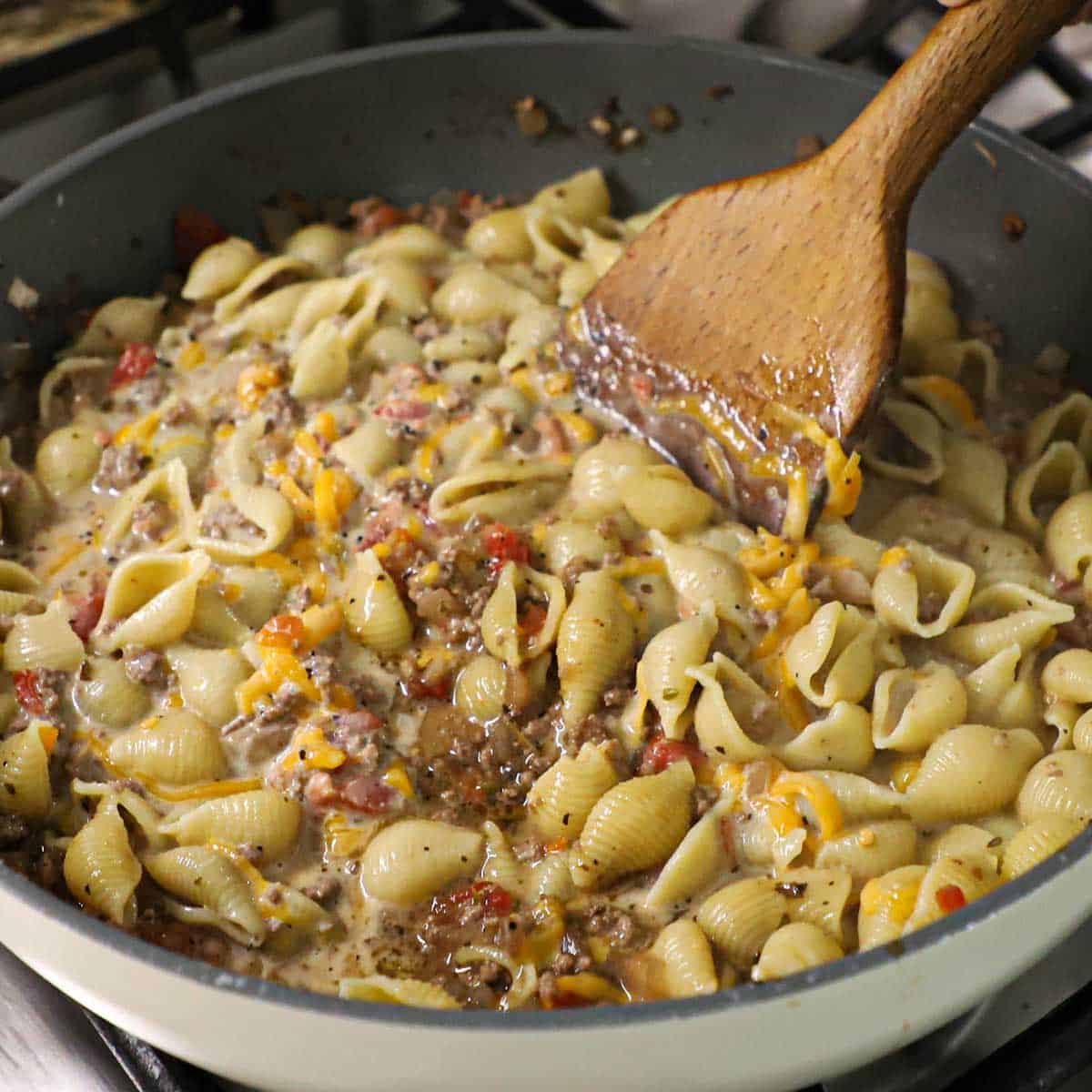 A person using a wooden spatula to stir a skillet filled with creamy beef and shells with shredded cheddar cheese beginning to melt as it is being stirred.
