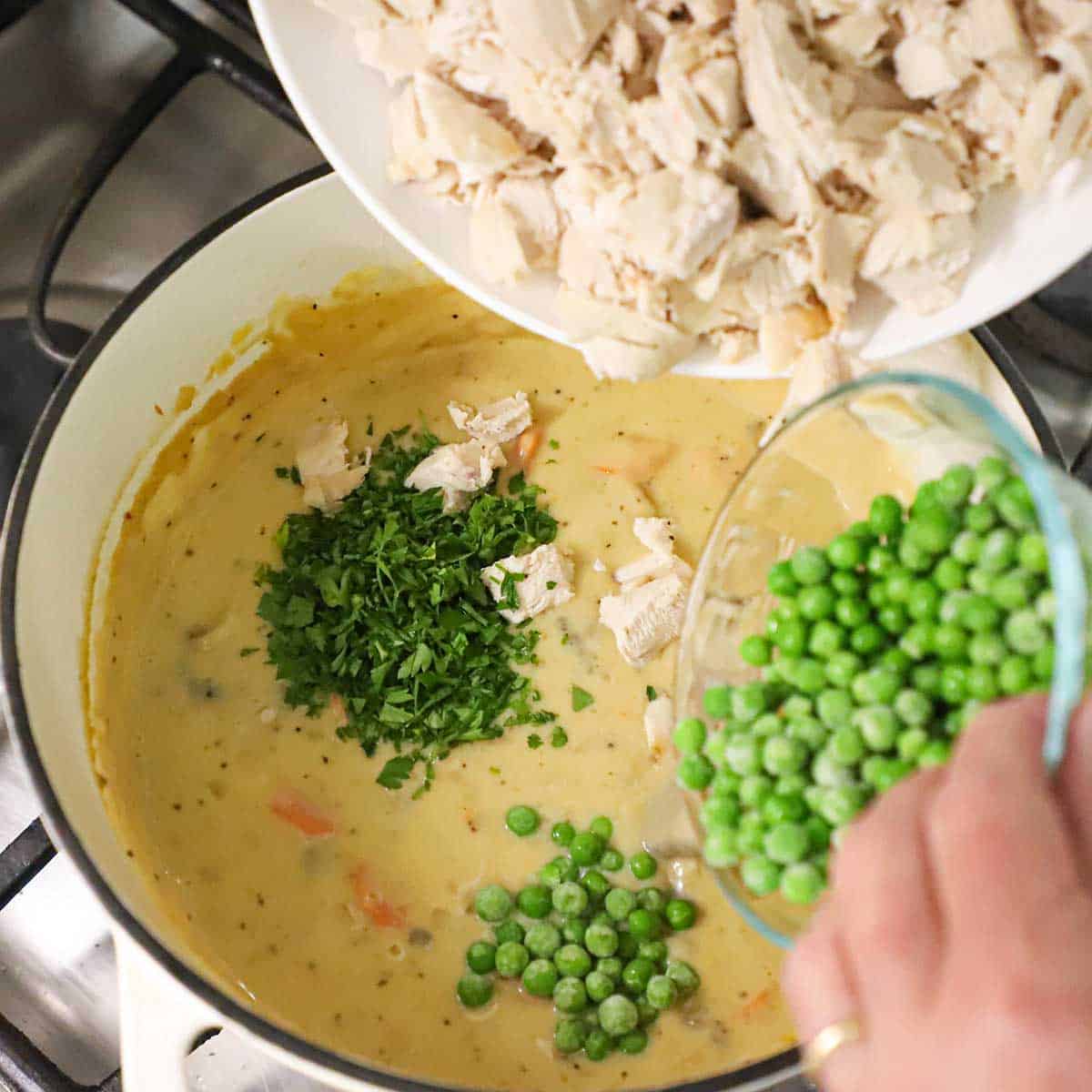 A person transferring cooked pieces of chicken from a bowl in one hand and another hand transferring peas into a pot filled with a simmering chicken pot pie filling.