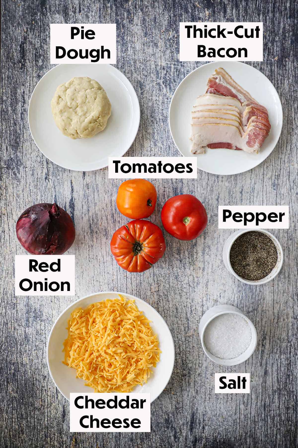 An arrangement of ingredients for tomato cheddar and bacon pie on a wooden grey background including whole tomatoes, bacon strips, a red onion, cheddar cheese, pie dough, salt, and pepper.