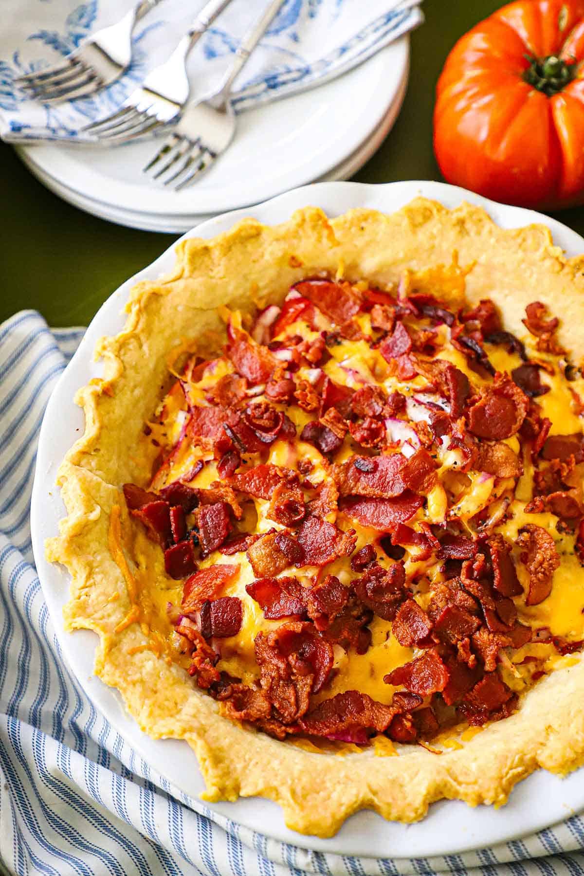 A freshly bake tomato cheddar and bacon pie sitting next to a blue and white napkin, plates with fork, and a whole tomato.