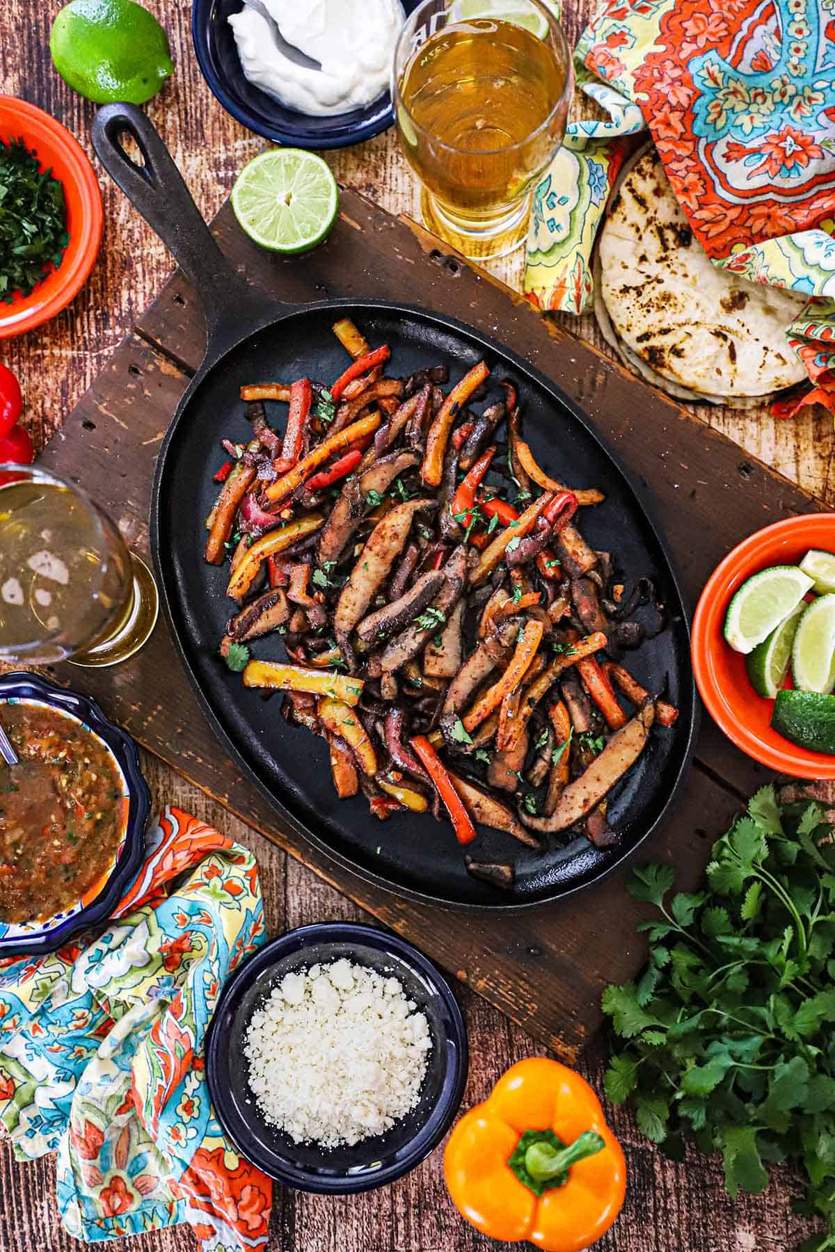 An overhead view of a fajita skillet filled with a serving of veggie fajitas with small bowls of cheese, limes, salsa, and cilantro surrounding the pan.