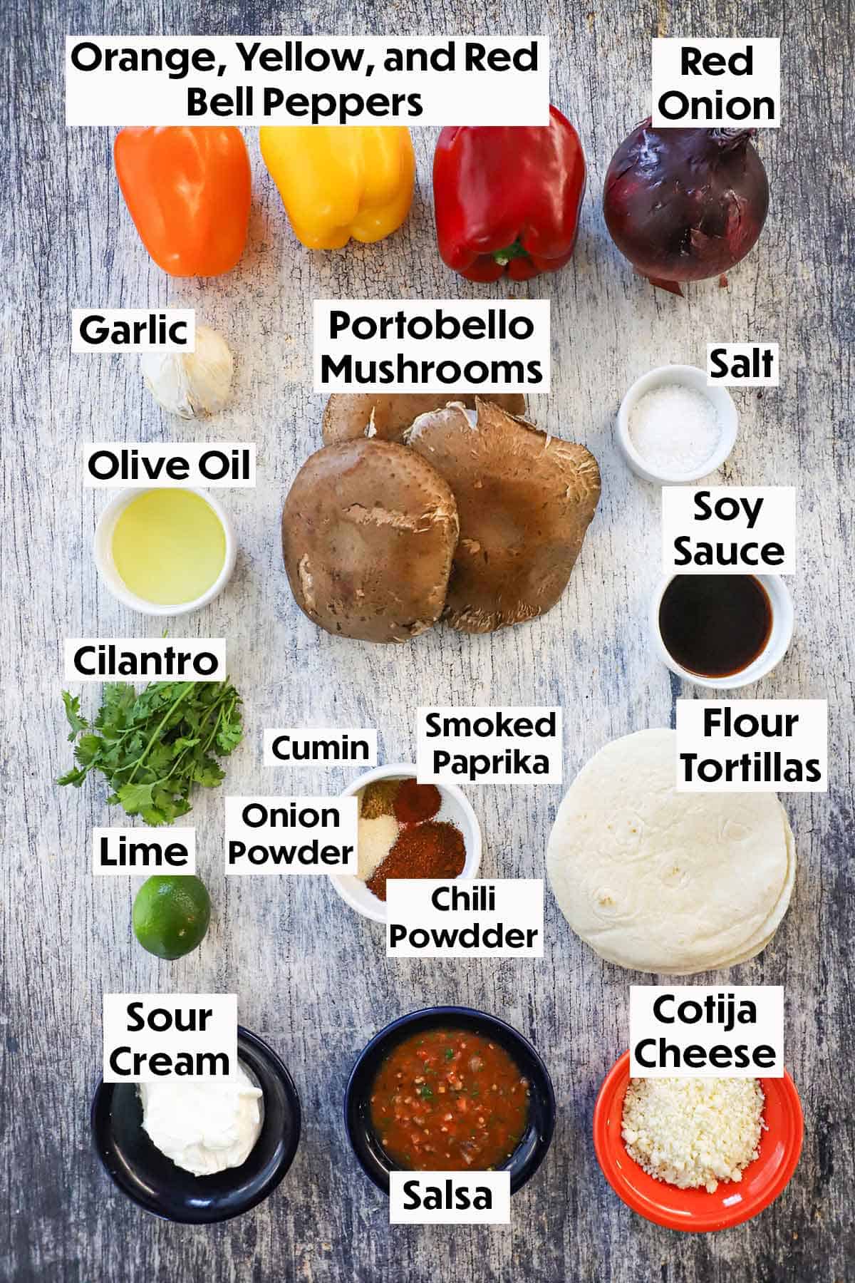 An arrangement of ingredients for veggie fajitas on a grey wooden background including portobello mushrooms, bell peppers, onion, garlic, oil, soy sauce, cilantro, seasonings, tortillas, sour cream, salsa, and cheese.