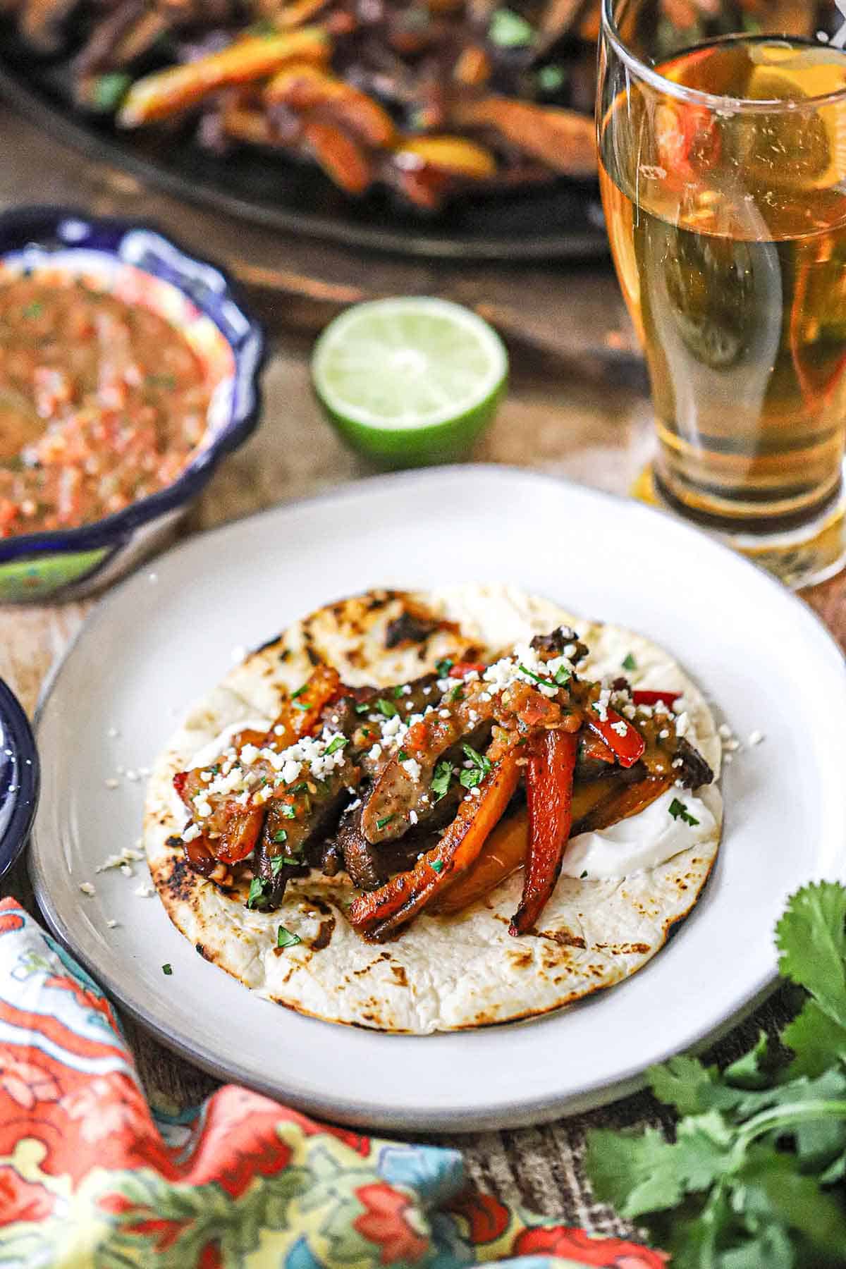 A white dinner plate filled with an open-face veggie fajita with a glass of beef nearby.