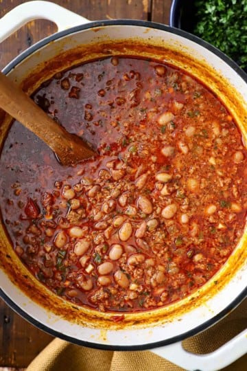 An overhead view of a large white pot filled with chorizo and bean soup with a wooden spoon in the middle of the soup.
