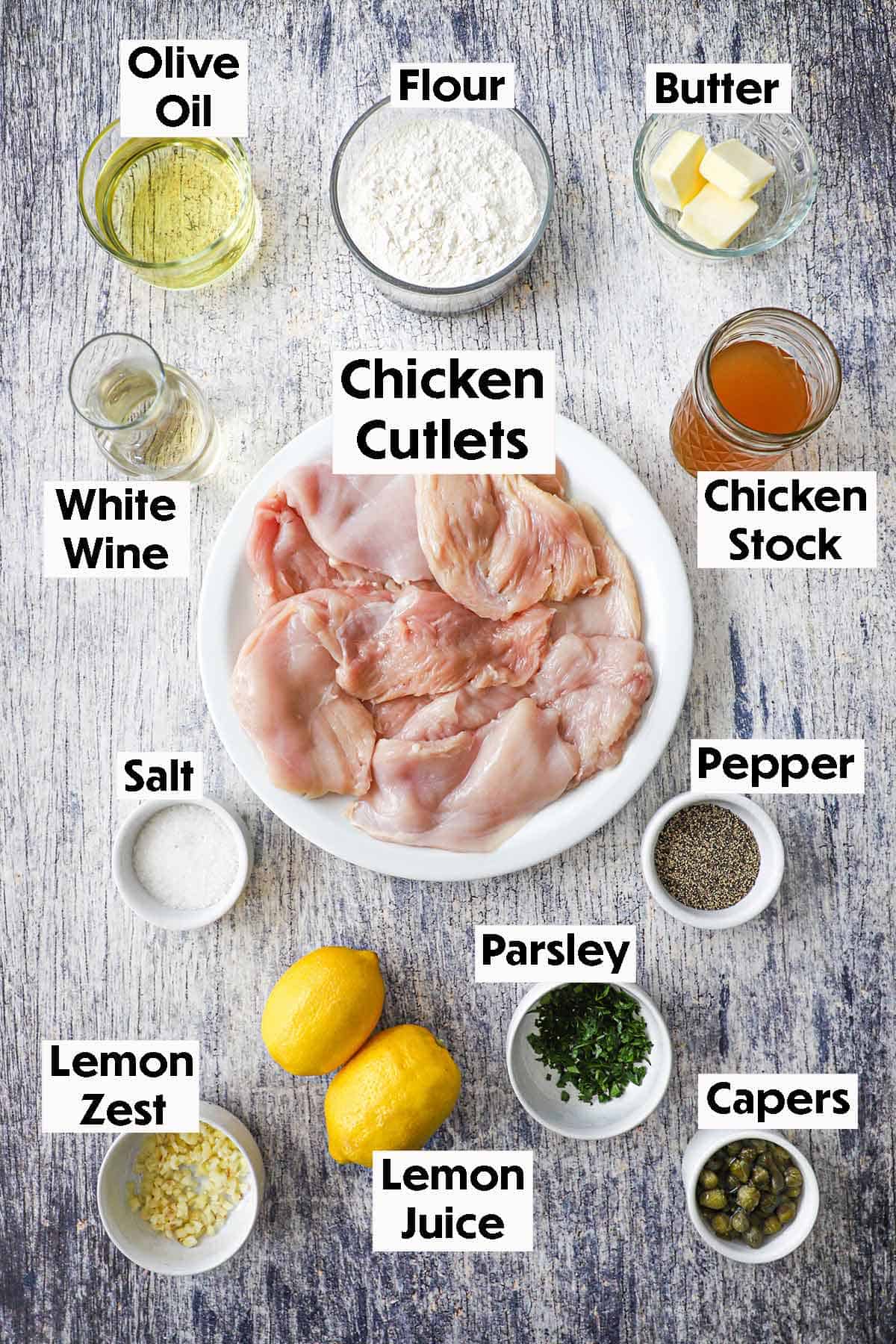 An arrangement of ingredients for homemade chicken piccata on a grey wooden background including thin chicken breast cutlets, olive oil, flour butter, wine, chicken stock, seasonings, lemons, parsley, and capers.