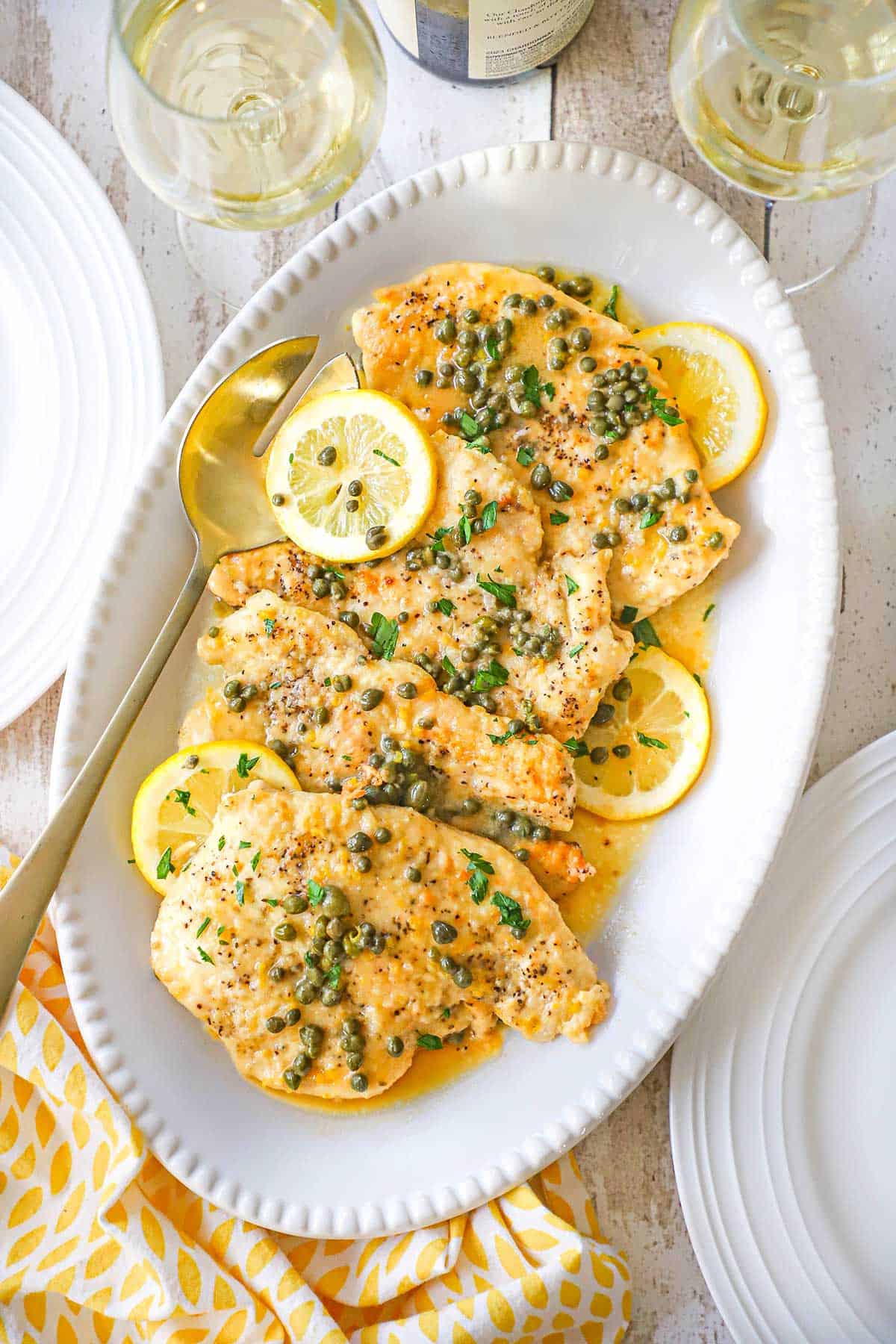 An overhead view of a beaded white platter filled with a row of freshly prepared classic chicken piccata cutlets with lemon wedges placed along the side of the dish.