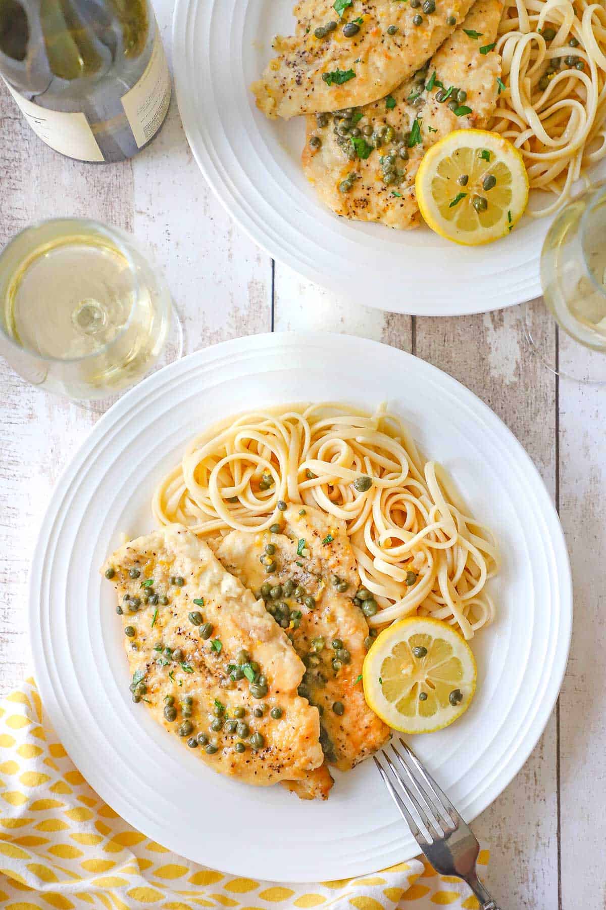 Two white dinner plates filled with servings of homemade chicken piccata and linguine with two glasses of white wine near the plates.