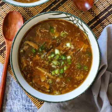 An overhead view of a festive soup bowl filled with homemade Chinese sweet and sour soup with a wooden spoon nearby and is garnished with chopped scallions.