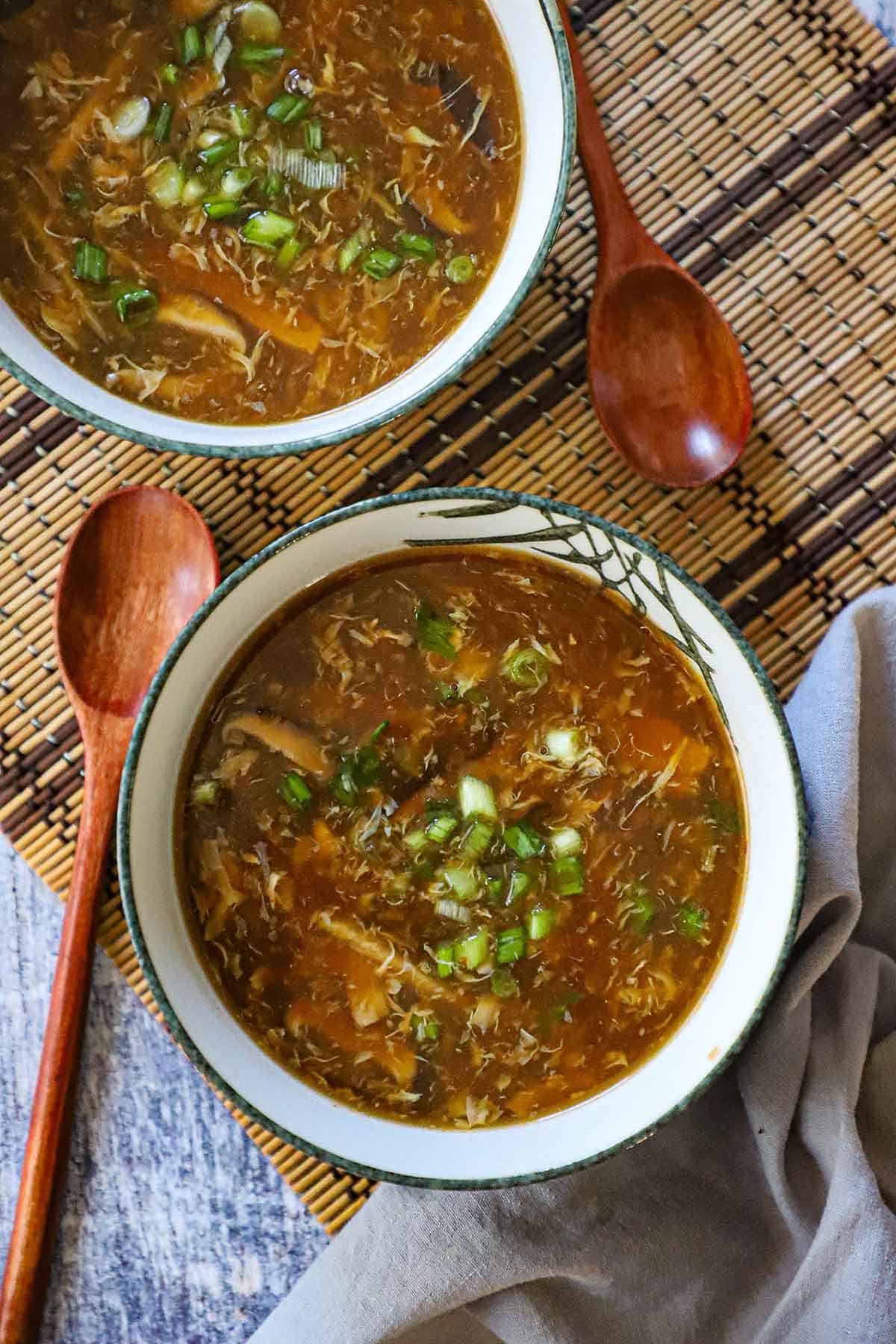 An overhead view of two bowls on a wicker placemat and that are filled with servings of homemade Chinese sweet and sour soup with wooden spoons flanking each bowl.