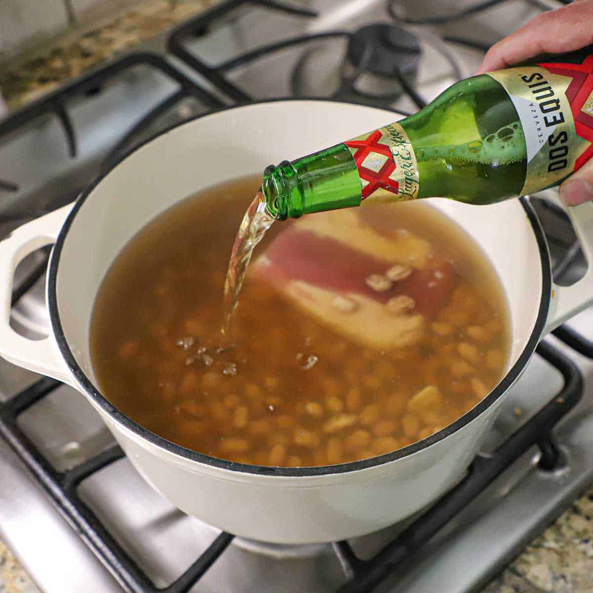 A person pouring beer from a bottle into a pot that is filled with dried pinto beans, water, chicken stock, and a salt pork.