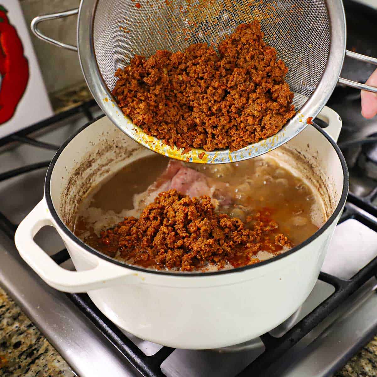 A person dumping cooked crumbled chorizo sauce from a sieve into a pot filled with simmering broth, a salt pork, and pinto beans.