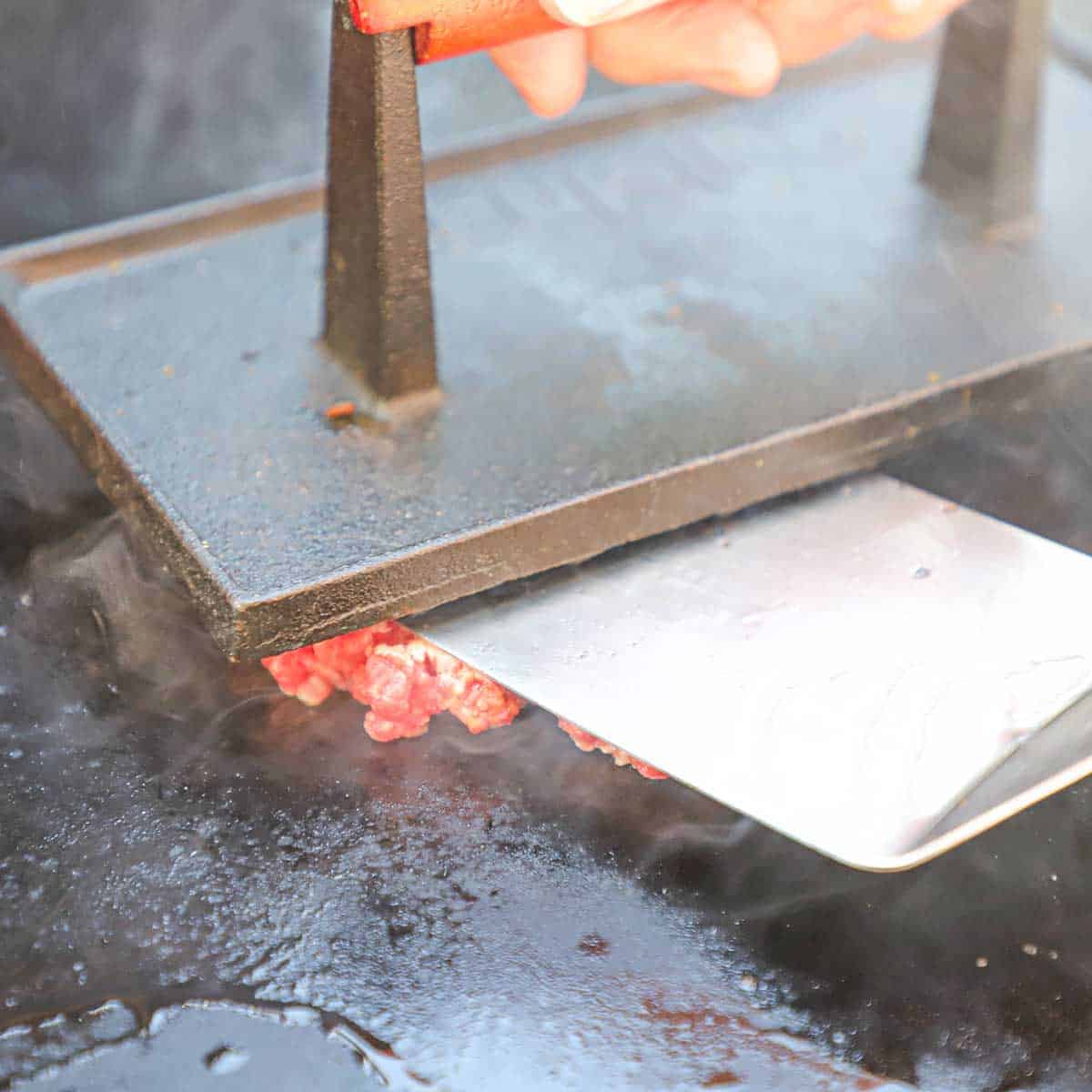 A person using a hamburger smasher and a large spatula to flatten ground hamburger on a hot griddle.