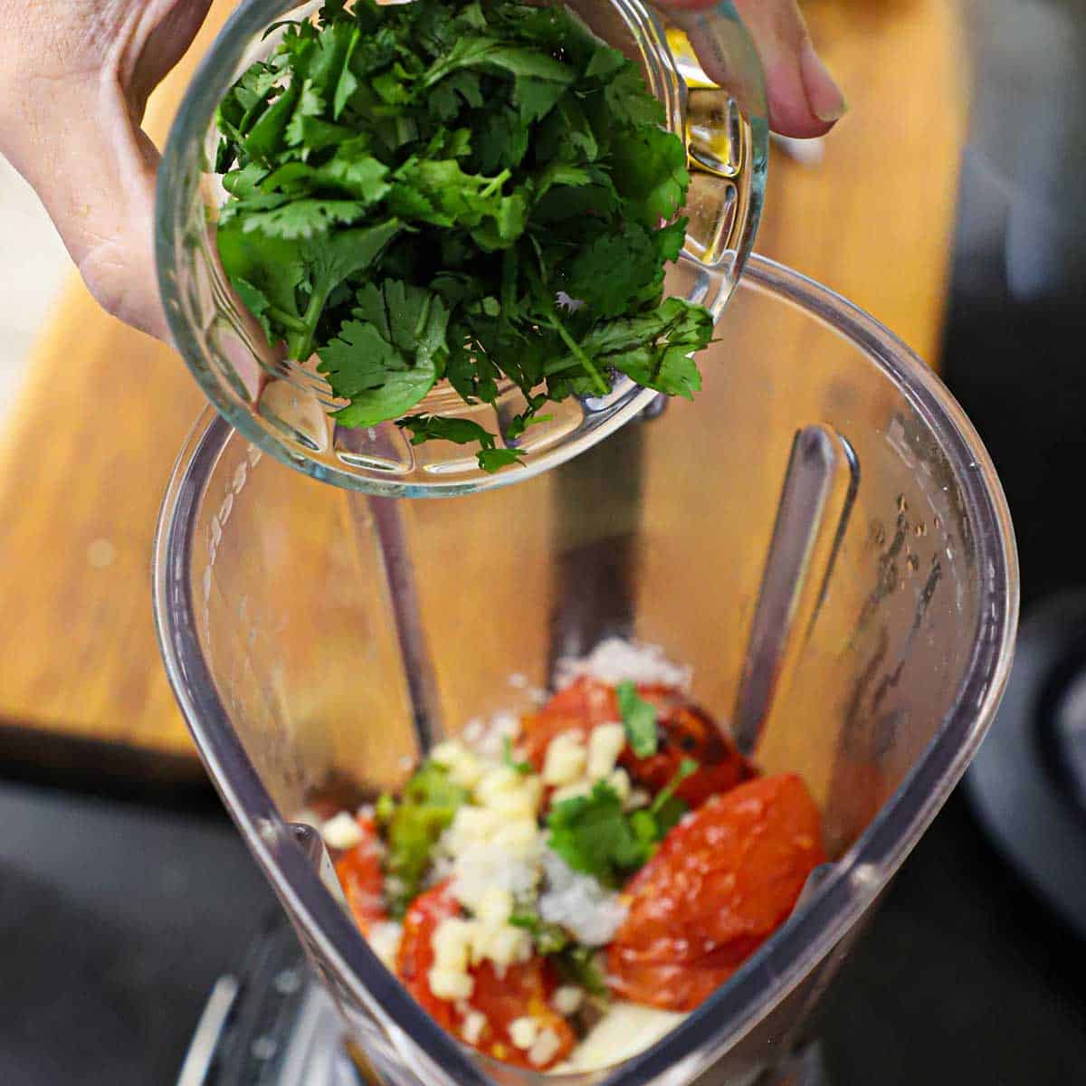 A person transferring chopped cilantro into a blender that is filled with roasted tomatoes, onions, peppers, and garlic.