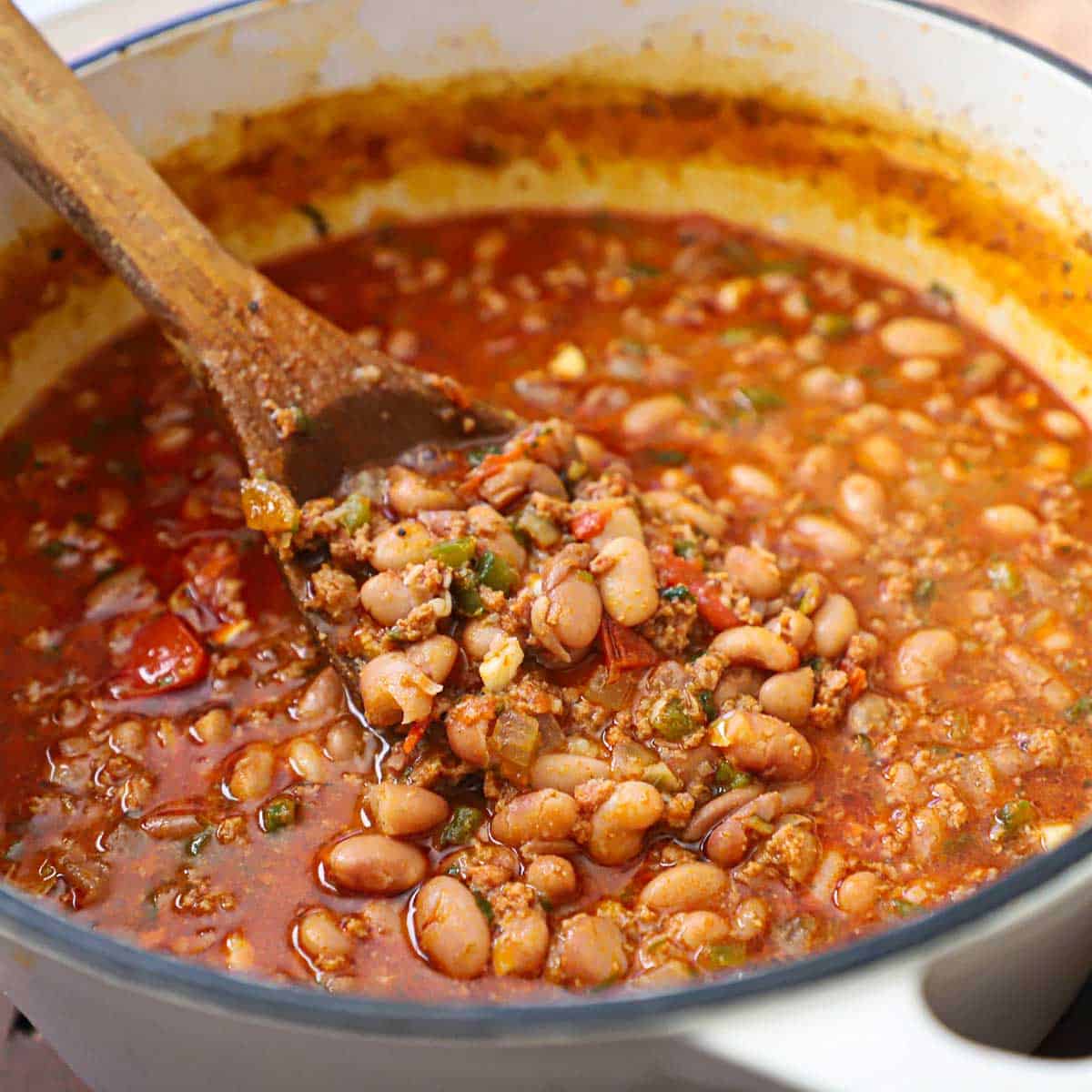 A person using a wooden spoon to stir a white pot of simmering chorizo and bean soup.