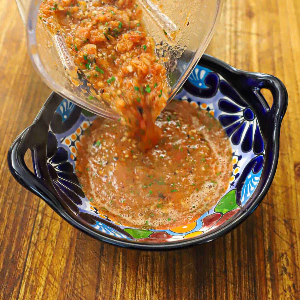 A person pouring roasted tomato salsa from a blender into a festive salsa bowl that is resting on a wooden cutting board.