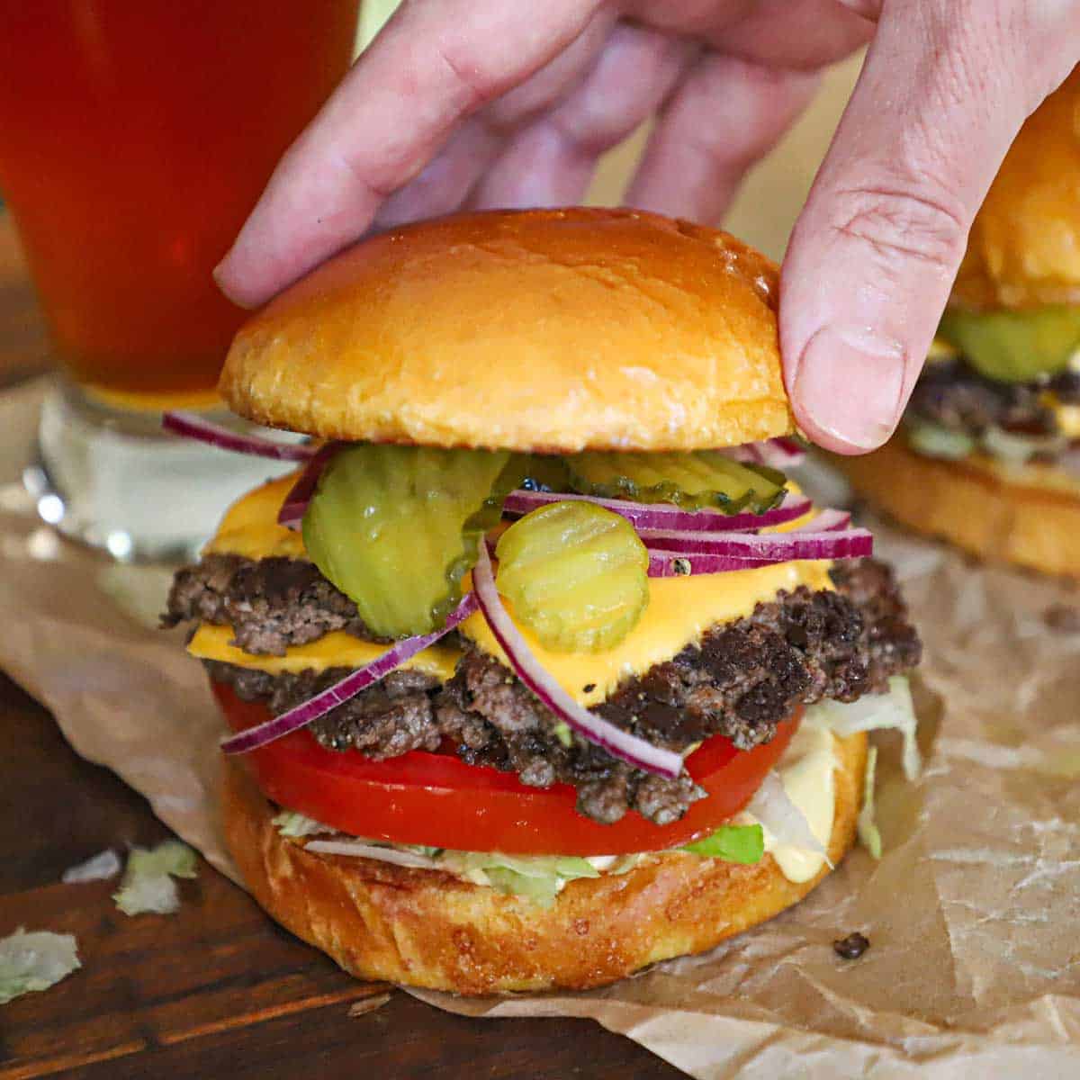 A person placing the top half of a toasted brioche hamburger on top of a double-stacked smash burger with American cheese, pickles, onion, tomato, lettuce, and burger sauce.