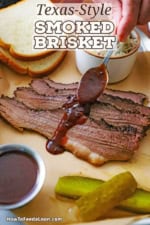 A person using a spoon to drizzle barbecue sauce over three strips of Texas-style smoked brisket on a platter next to sliced bread, barbecue sauce, and pickles.