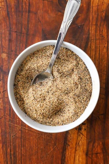 A small white bowl filled with Best Beef Rub with a spoon in the middle of it while the bowl is resting on a wooden cutting board.